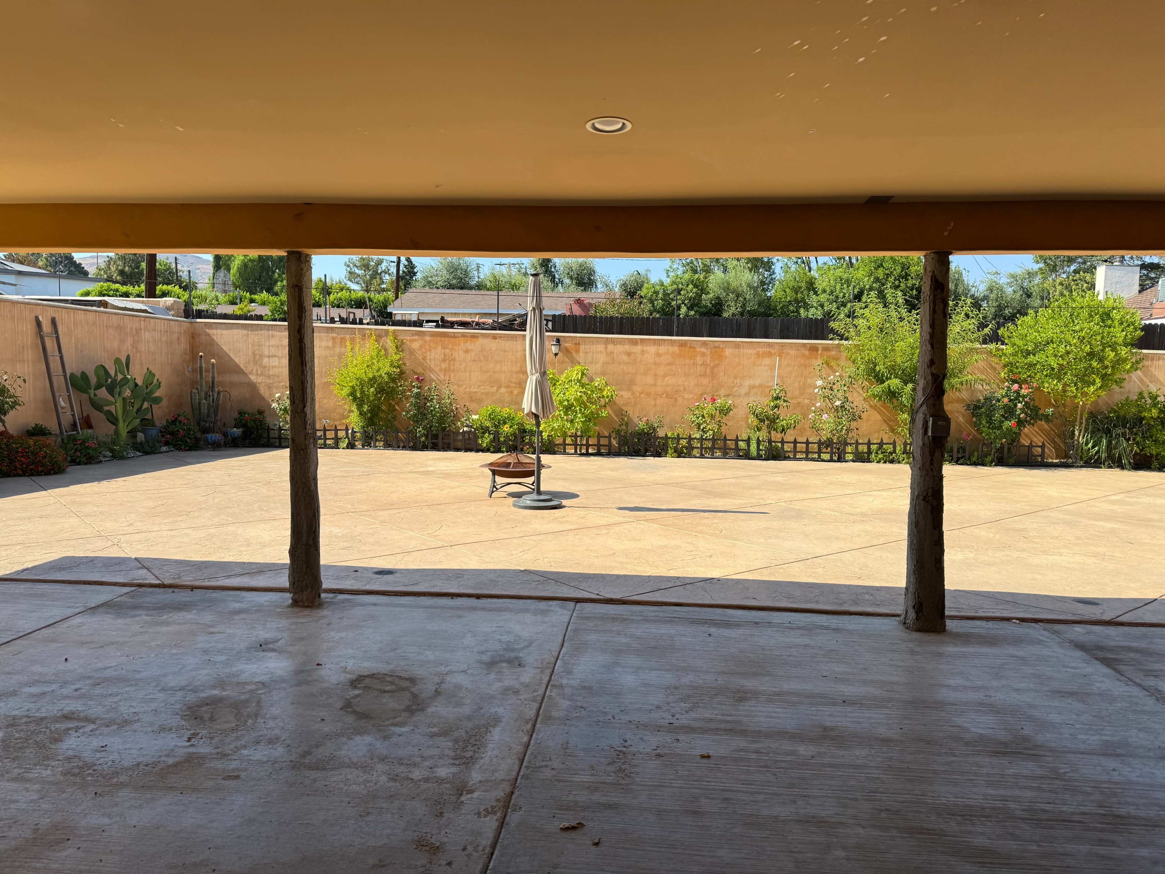 The image shows a spacious outdoor area with a concrete floor, surrounded by greenery and a few potted plants, viewed from underneath a sheltered structure.