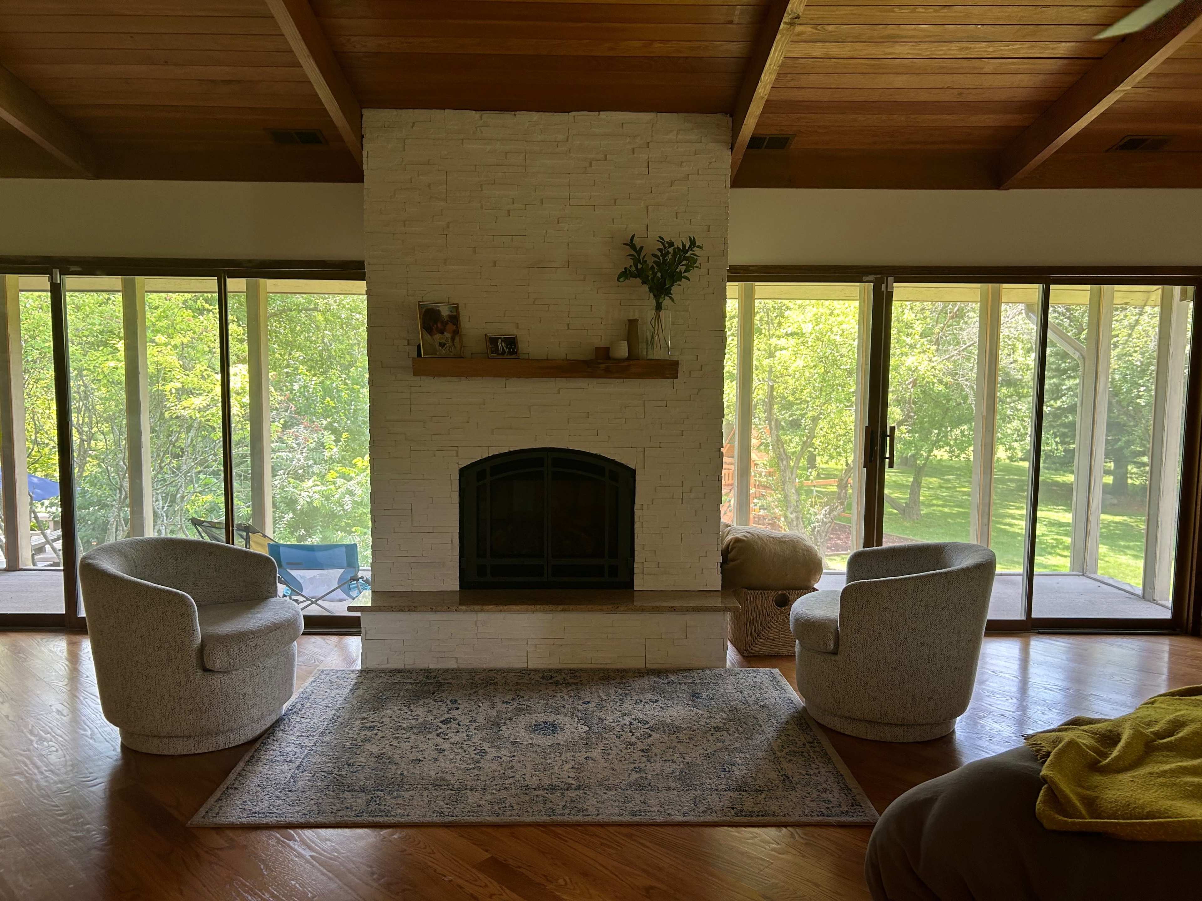 The living room features a white stone fireplace with two upholstered chairs on either side and large glass doors opening to a green outdoor space.