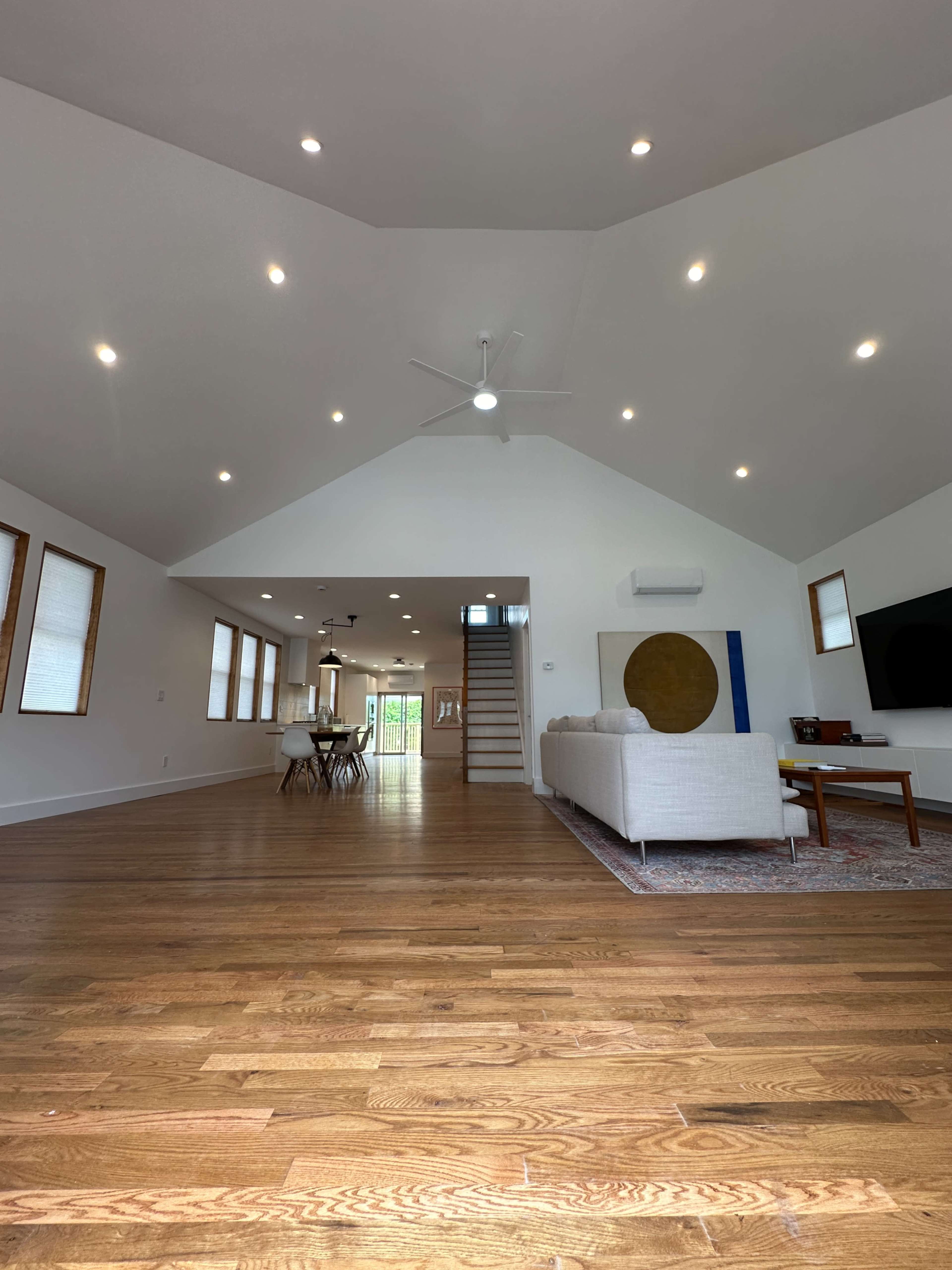 The image depicts a spacious, modern living room with hardwood floors, a white sofa, and large windows, leading to a staircase and dining area in the background.
