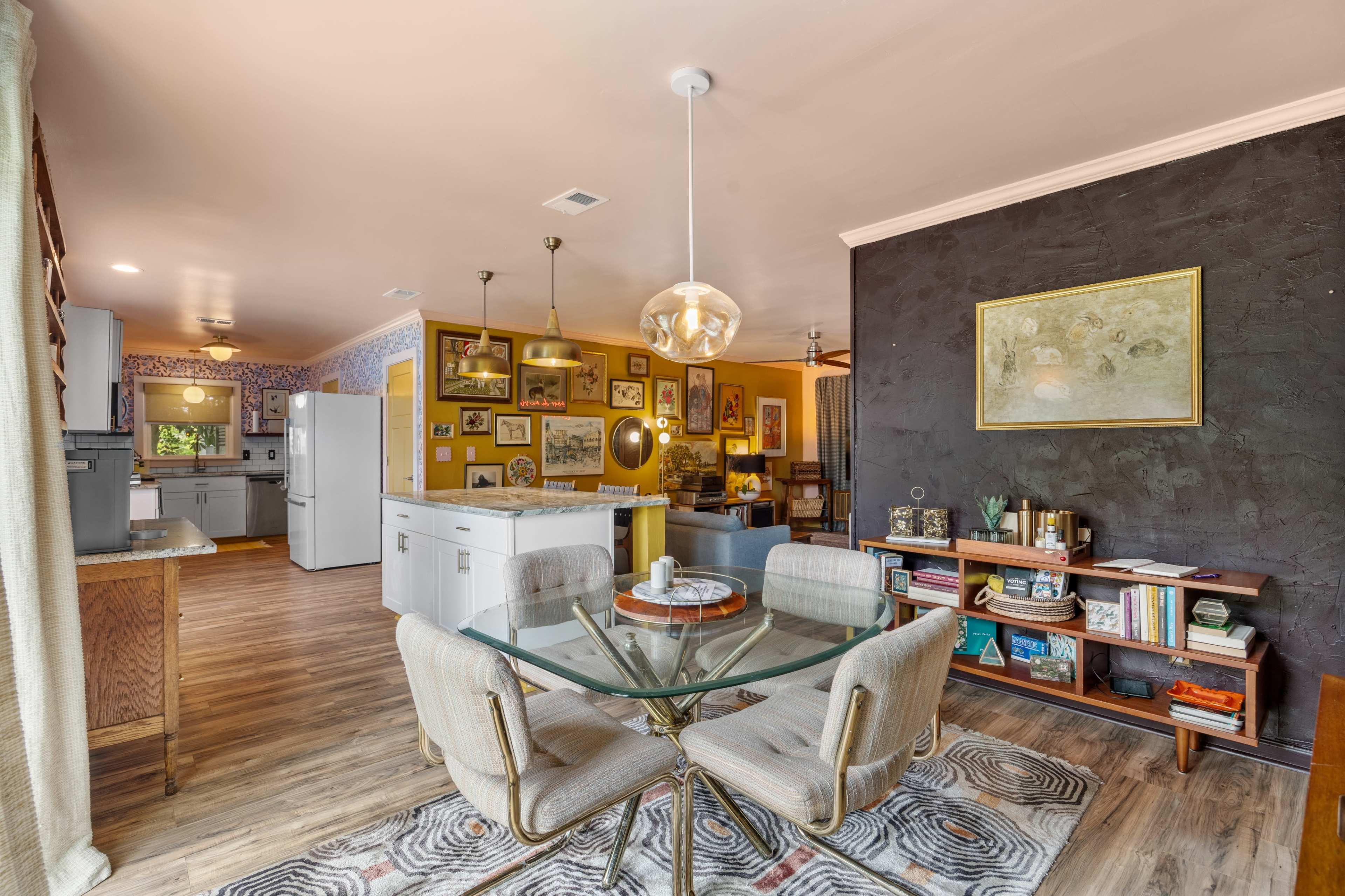 The image features a dining area with a glass table and chairs, a modern kitchen in the background, and walls adorned with artwork and decor.