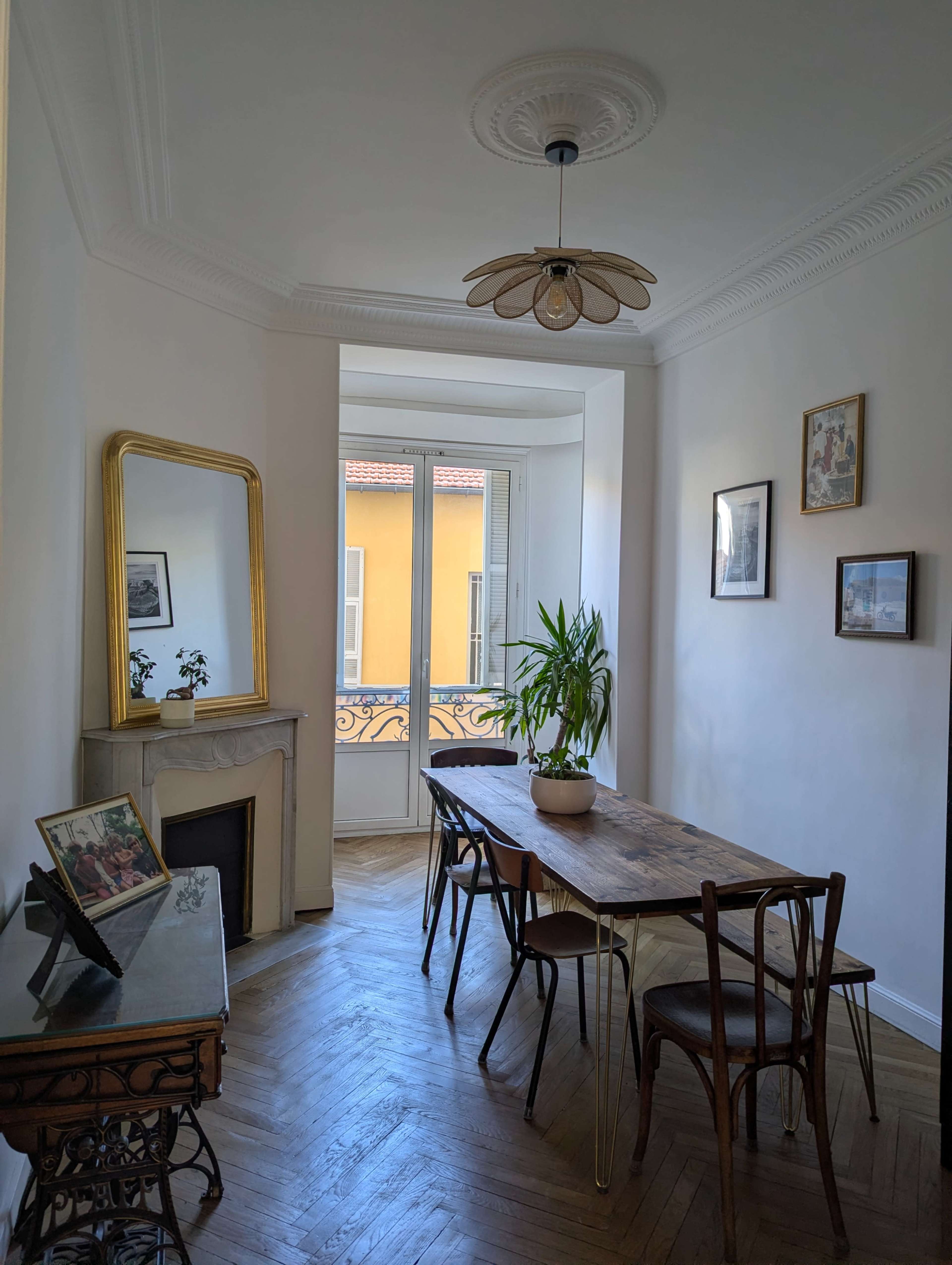 The image shows a bright dining area featuring a wooden table surrounded by chairs, a large mirror, a potted plant, and a view of a balcony through a glass door.