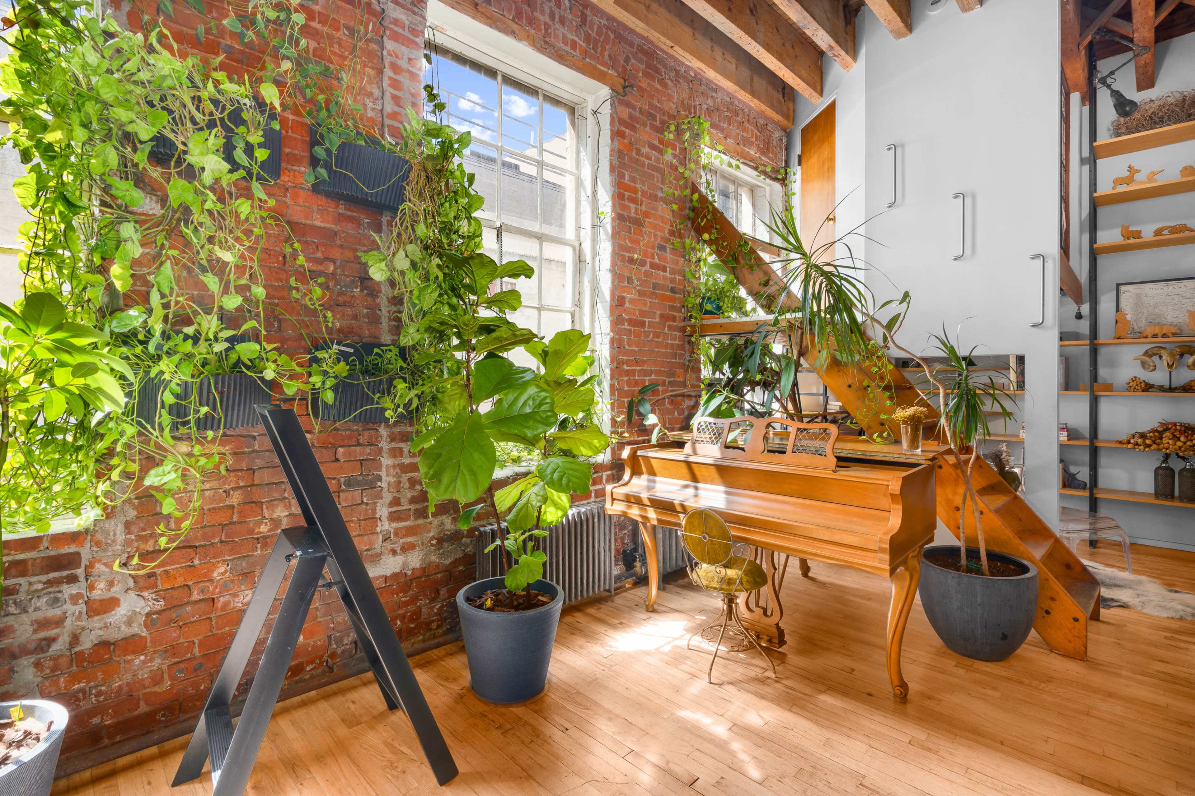 A wooden piano is positioned against a wall adorned with plants in a room featuring exposed brick and wooden beams.