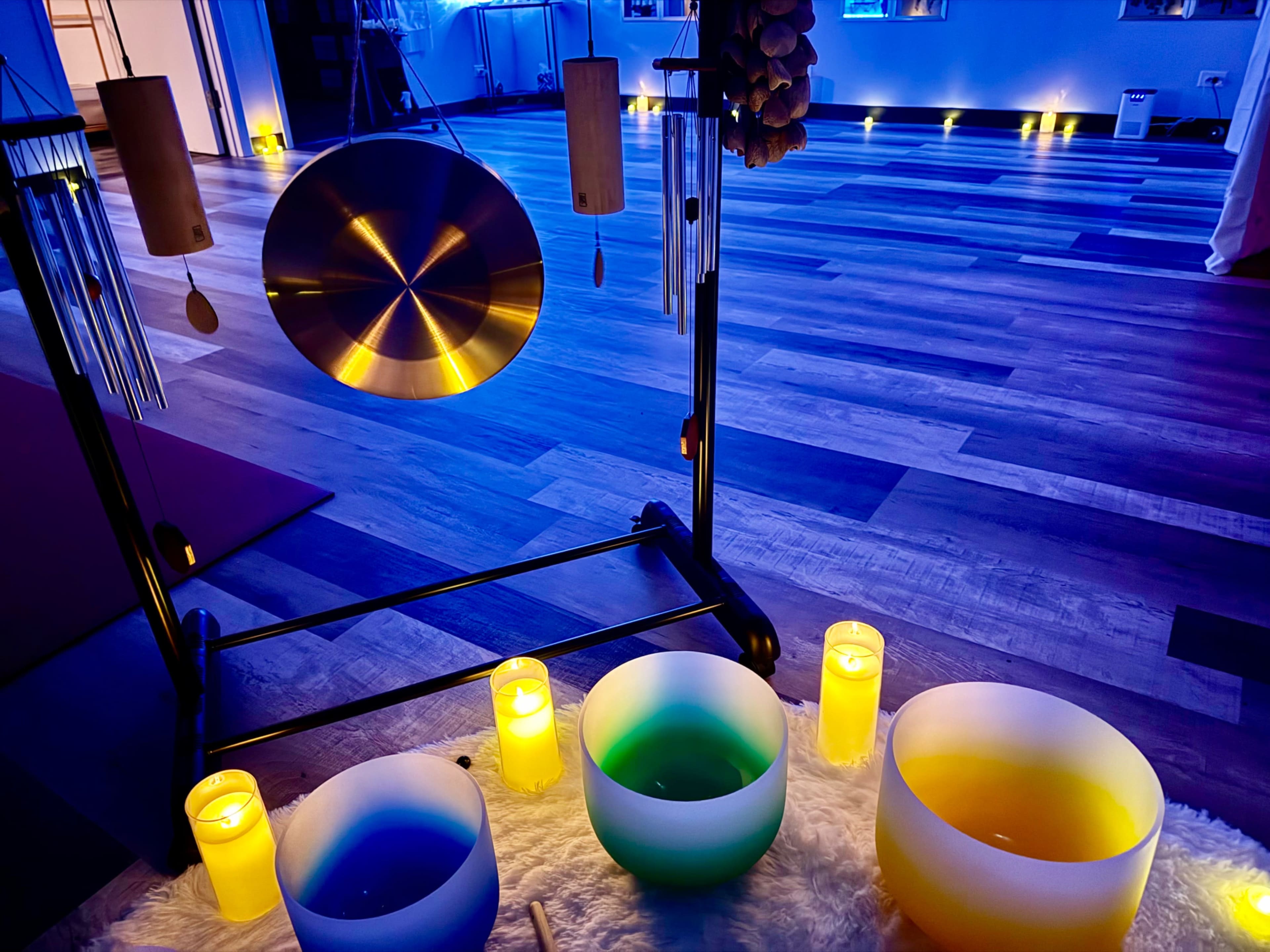 The image shows a sound therapy setup featuring a large gong, wind chimes, and three colored singing bowls, surrounded by lit candles on a softly illuminated wooden floor.