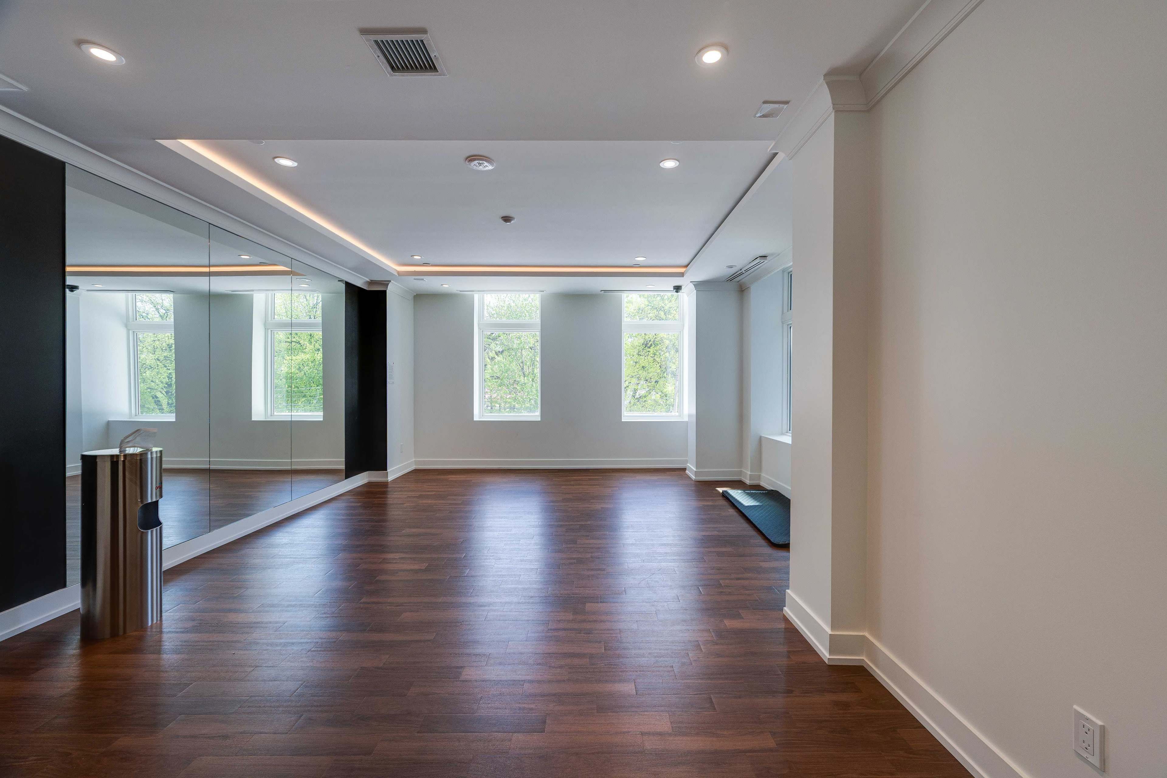 The image shows a spacious room with hardwood flooring, large windows, and a mirrored wall, creating an open and bright atmosphere.