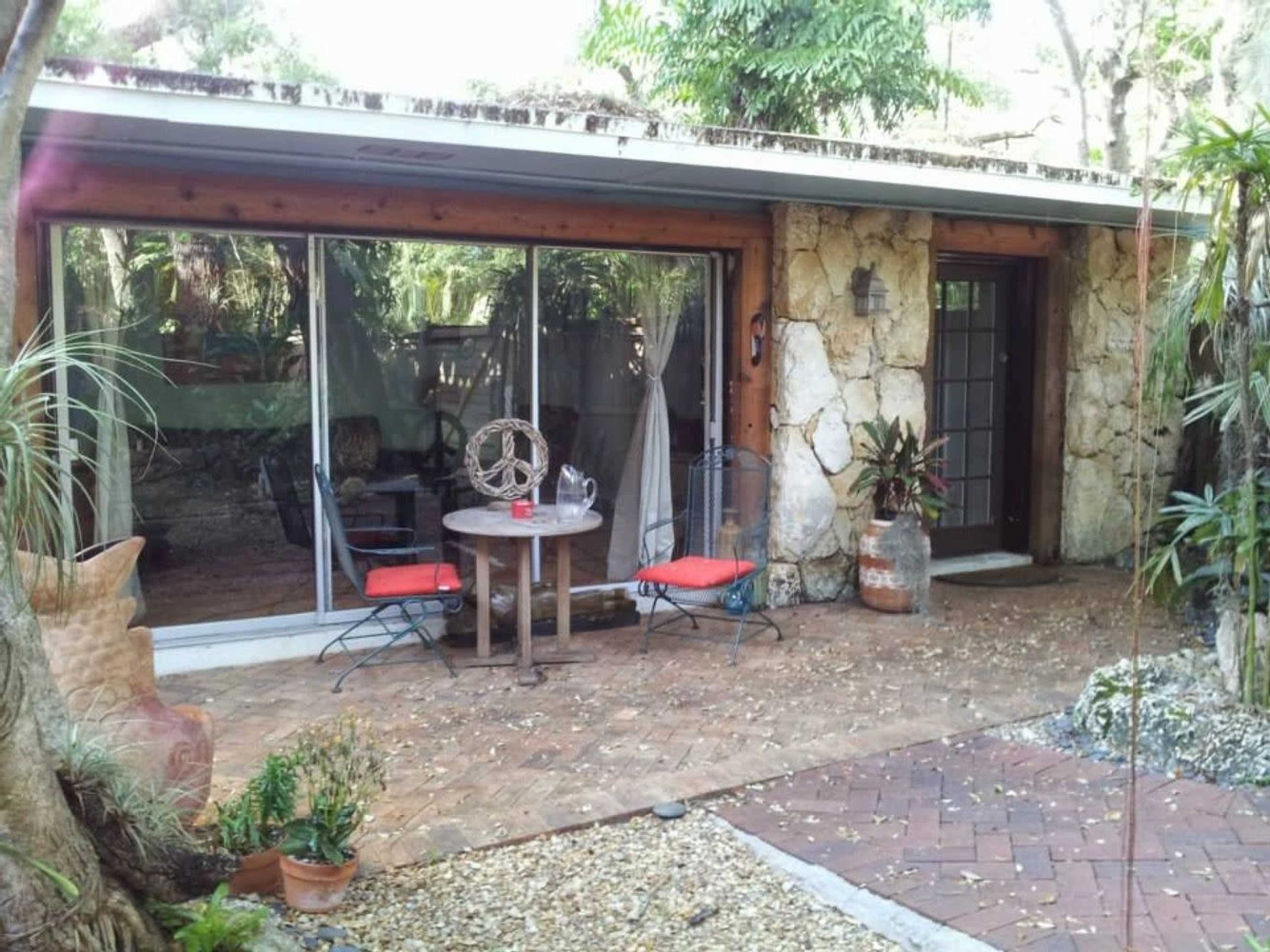 The image shows a small patio area with a round table and two chairs, surrounded by plants and a stone wall.
