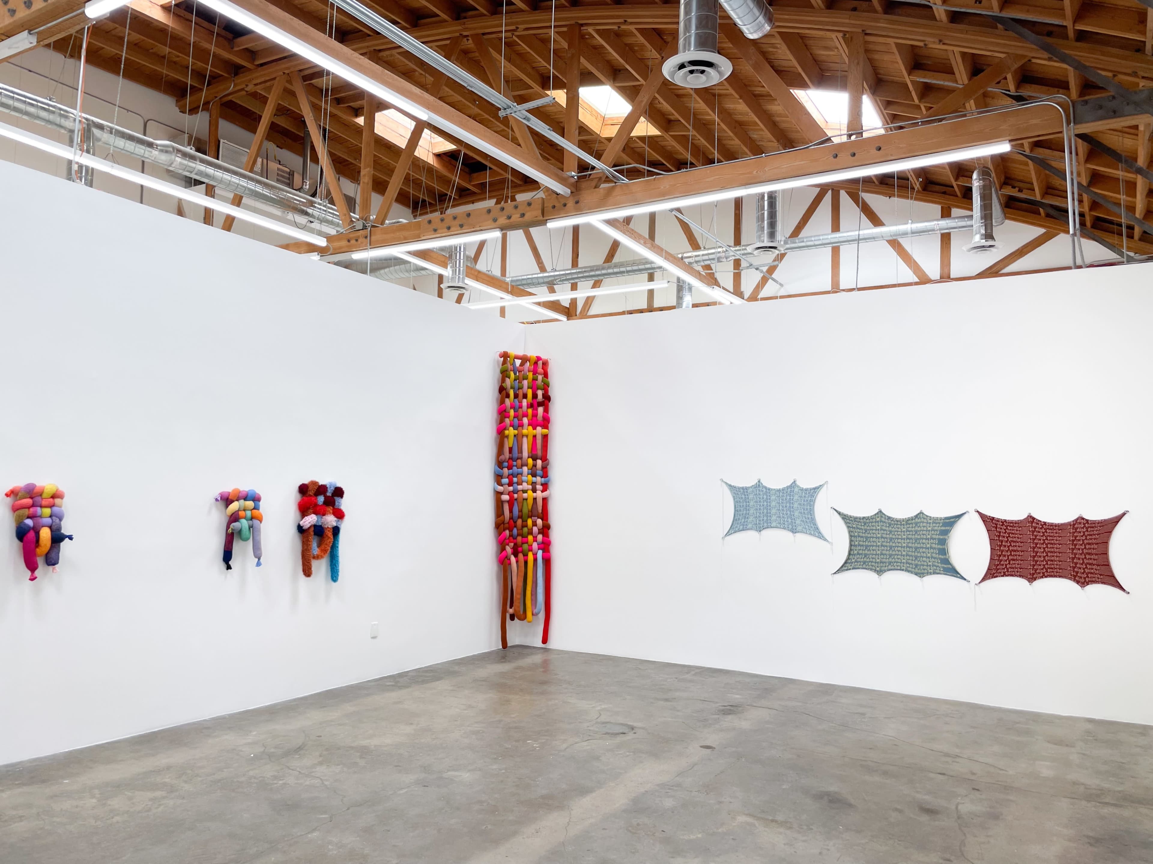The image shows an art gallery with colorful textile installations mounted on the white walls, featuring various shapes and patterns.