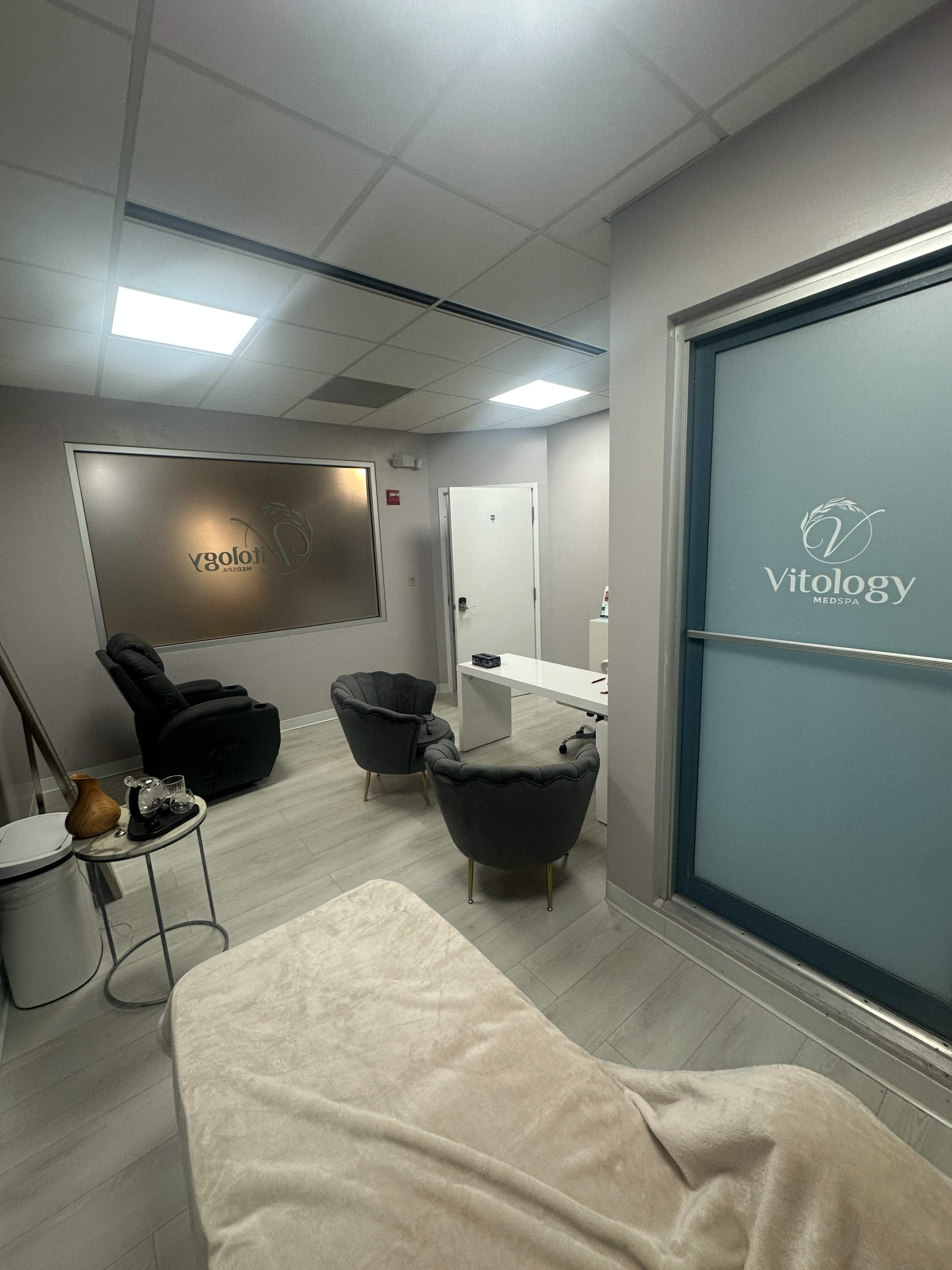 The image shows a sleek, modern waiting room with gray chairs, a small table, and a frosted glass door labeled "Vitology."