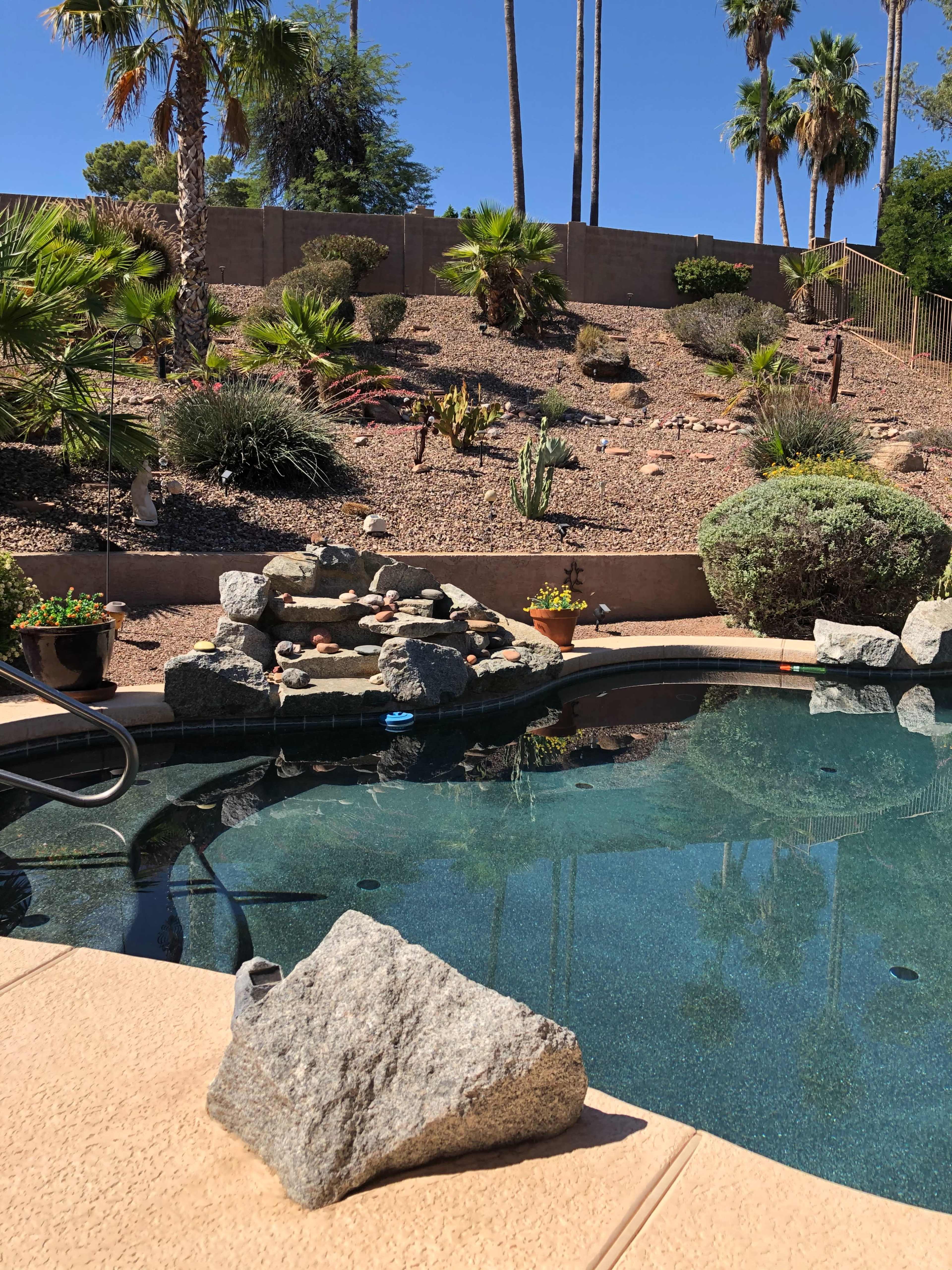 The image shows a swimming pool with a small rock waterfall surrounded by landscaped gardens and palm trees.