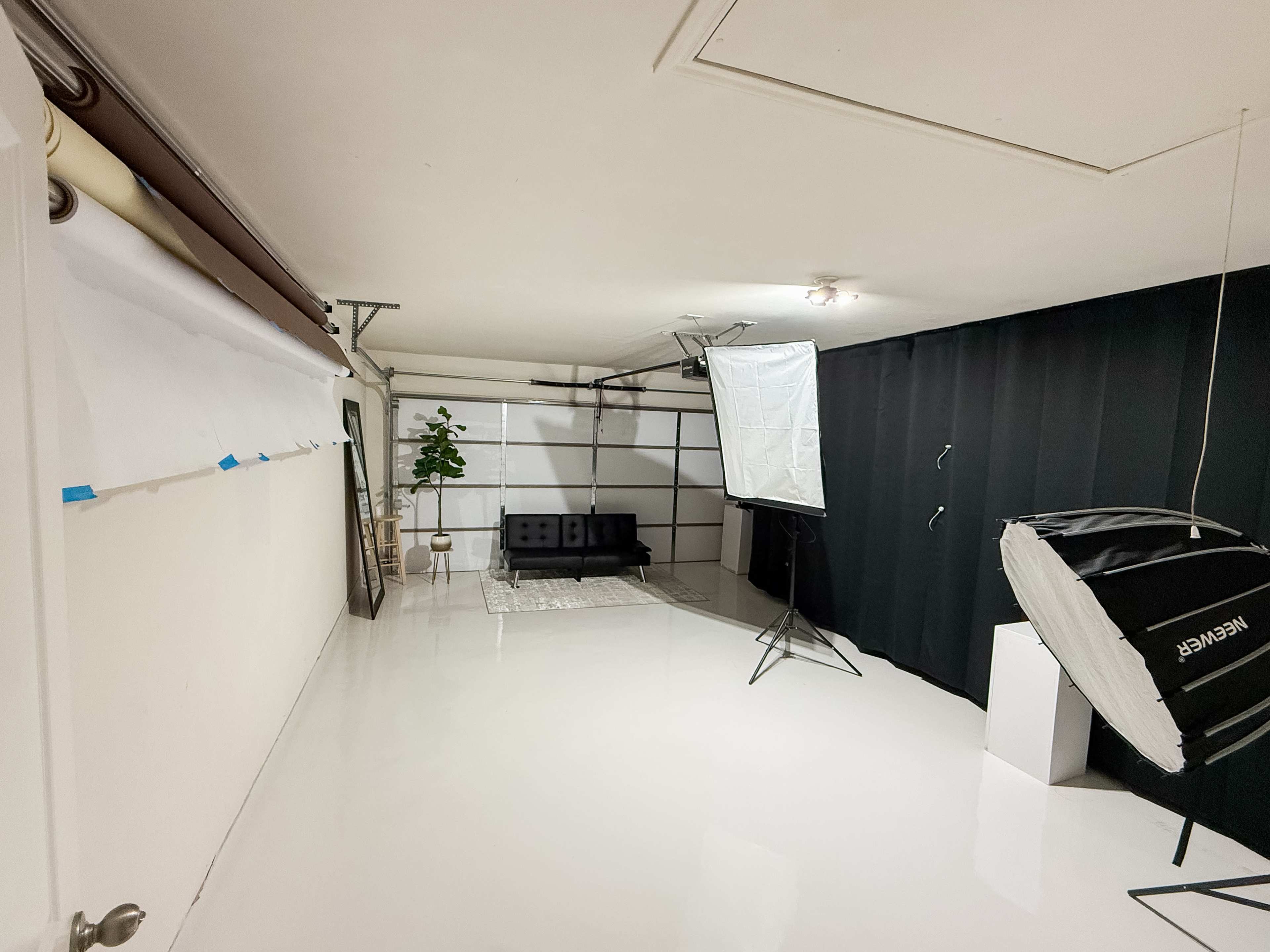 The image shows a minimalist photography studio with a black backdrop, a small black couch, a lighting setup, and a rolled backdrop at one end.