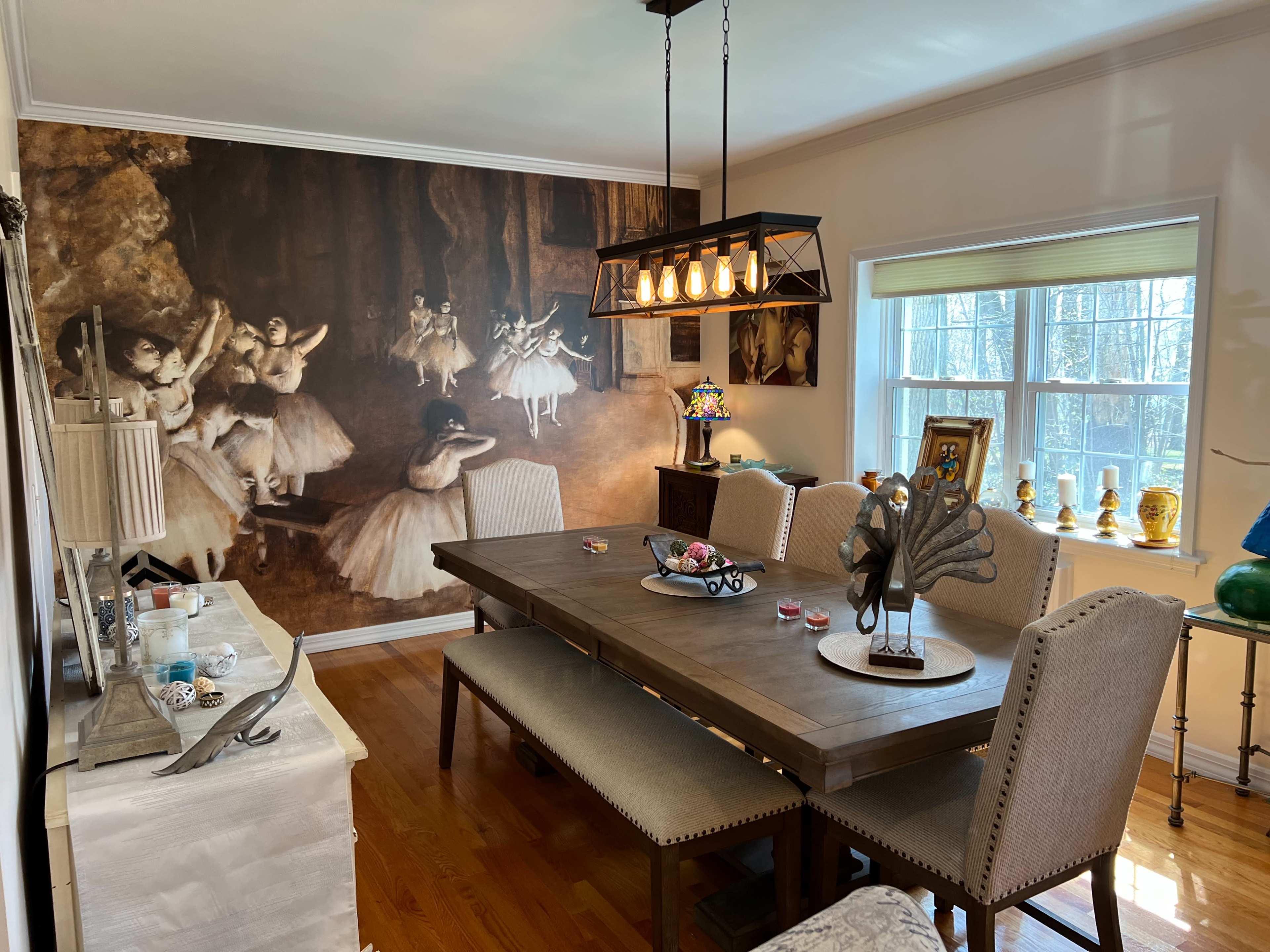 The dining room features a large wooden table surrounded by upholstered chairs, with a mural of ballet dancers on the wall and decorative items displayed on the sideboard and windowsill.