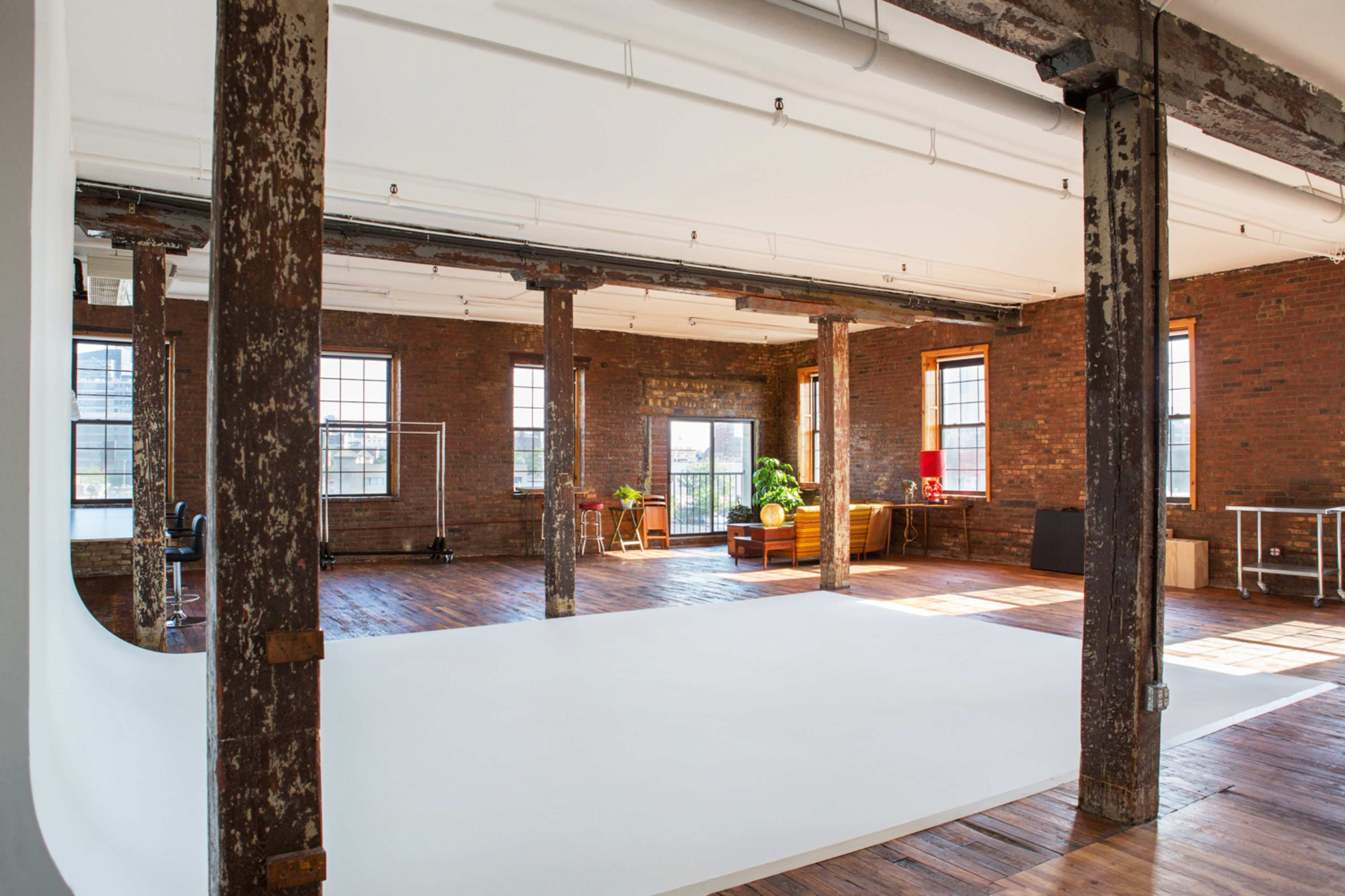 An industrial-style studio space with exposed brick walls, wooden floors, and large windows, featuring a white backdrop and several props for photography.