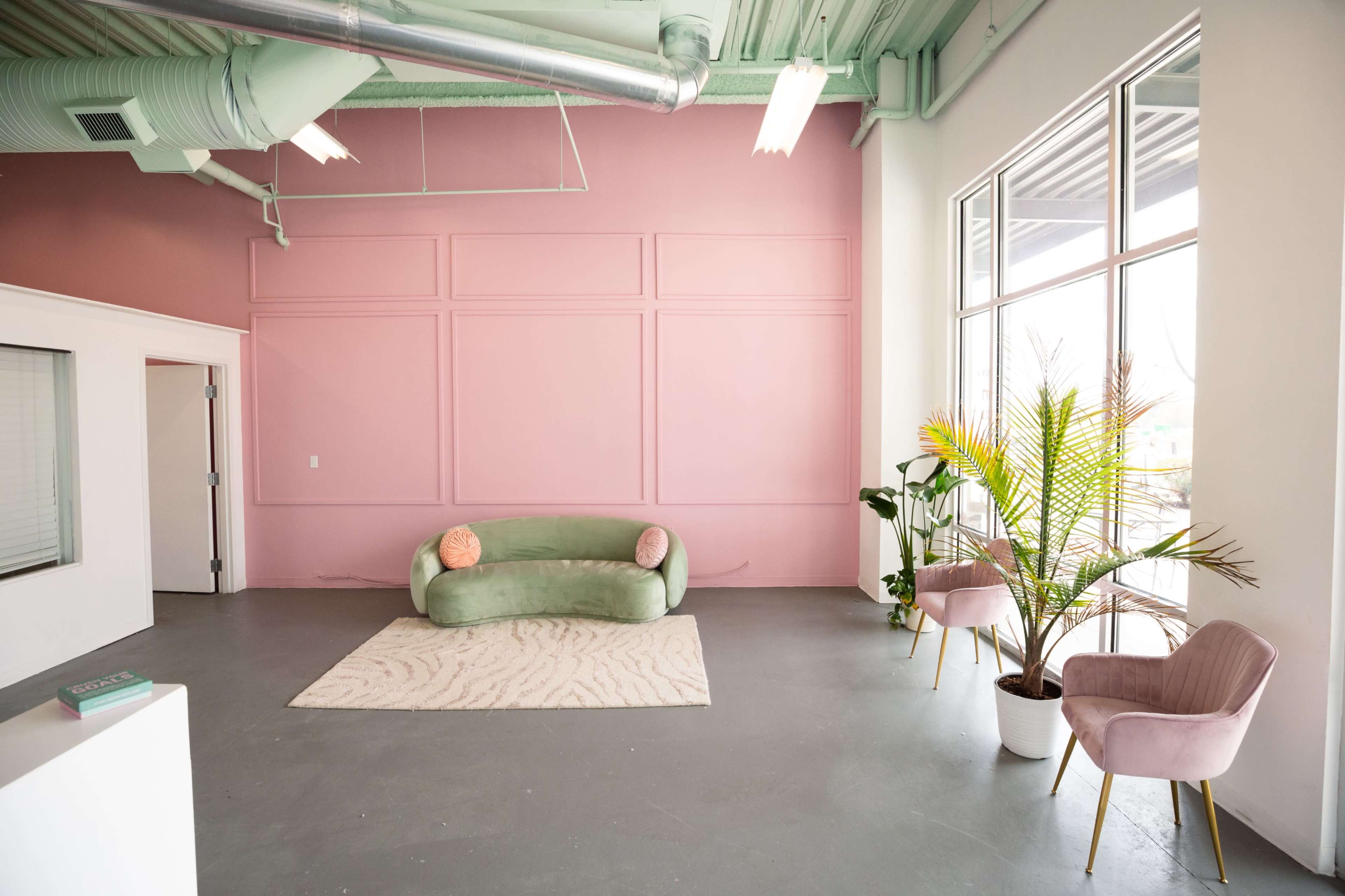 A spacious, minimalistic interior with a pink accent wall, a green sofa, two pink chairs, a rug, and a potted plant.