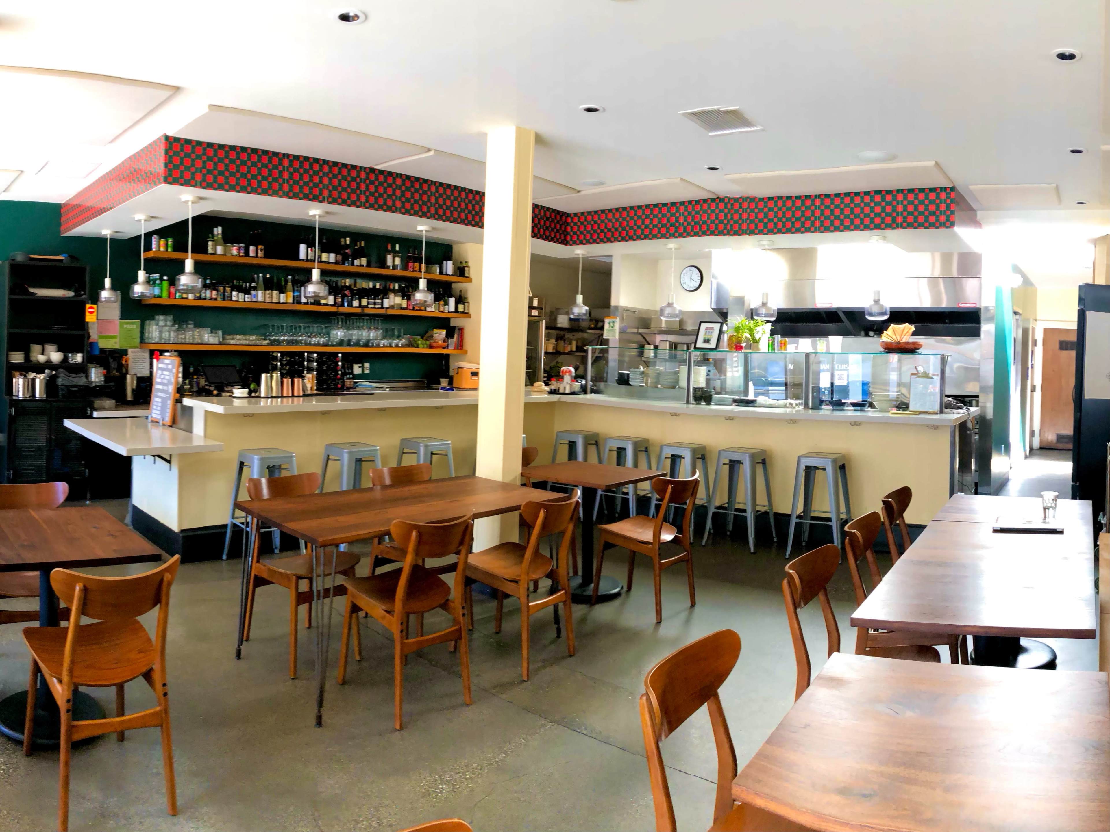 A spacious café features wooden tables, metal stools, and a counter with food preparation areas and shelves stocked with various items.