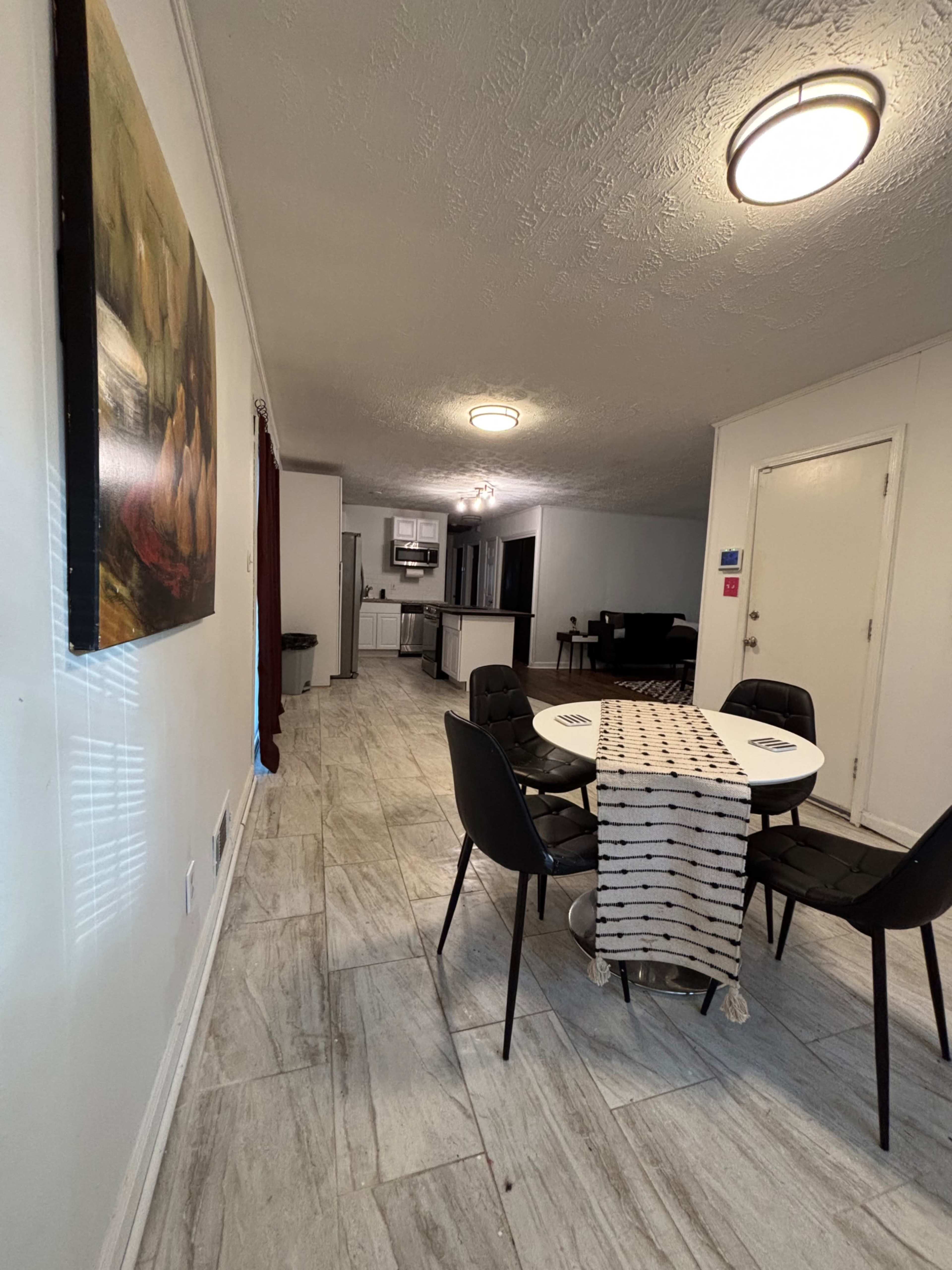 A dining area features a round table surrounded by black chairs, with a view of a kitchen and living space in the background.