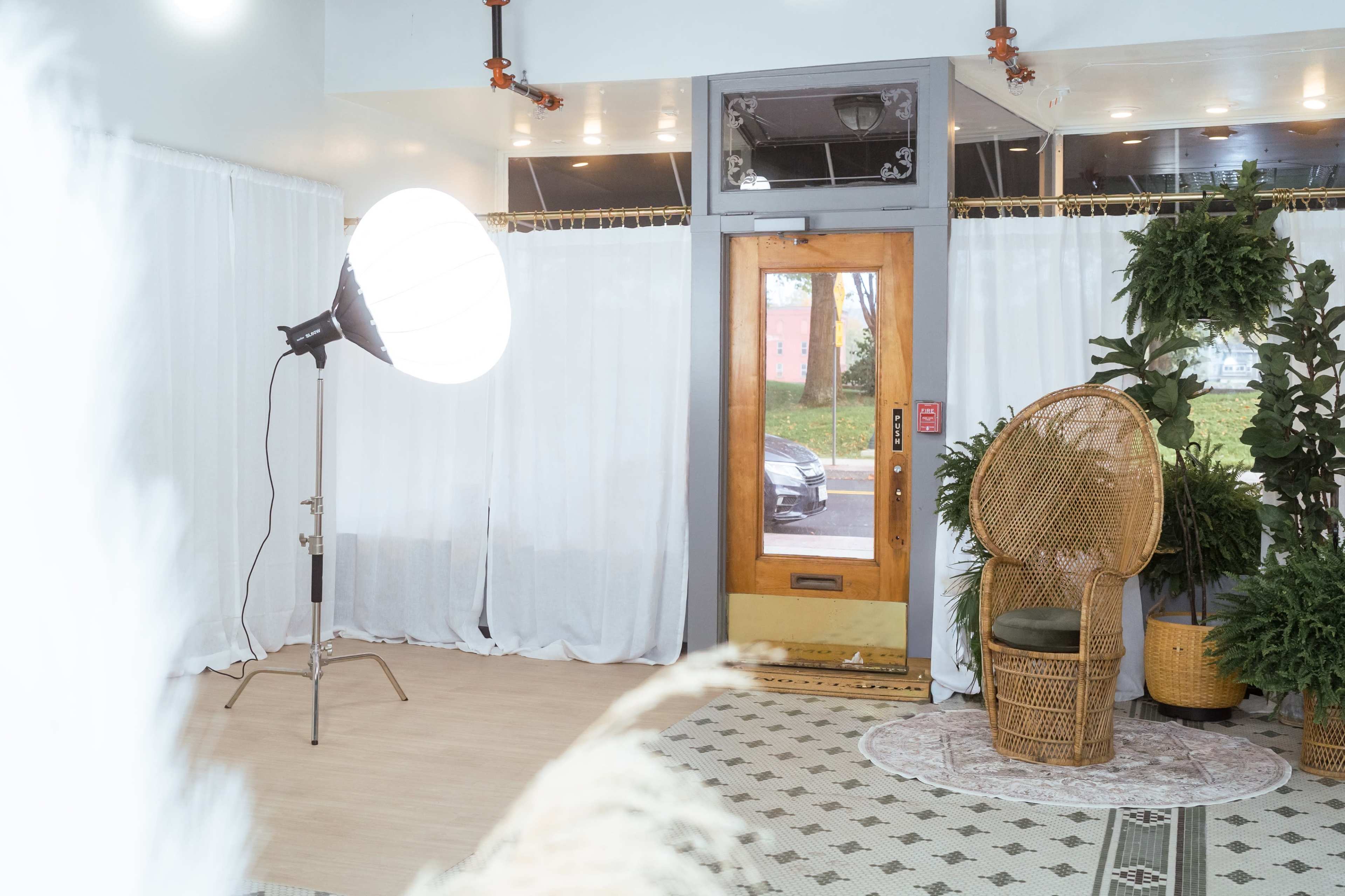 A large light fixture stands next to a wooden door, with a wicker chair positioned beside leafy plants in a room featuring white curtains and patterned flooring.