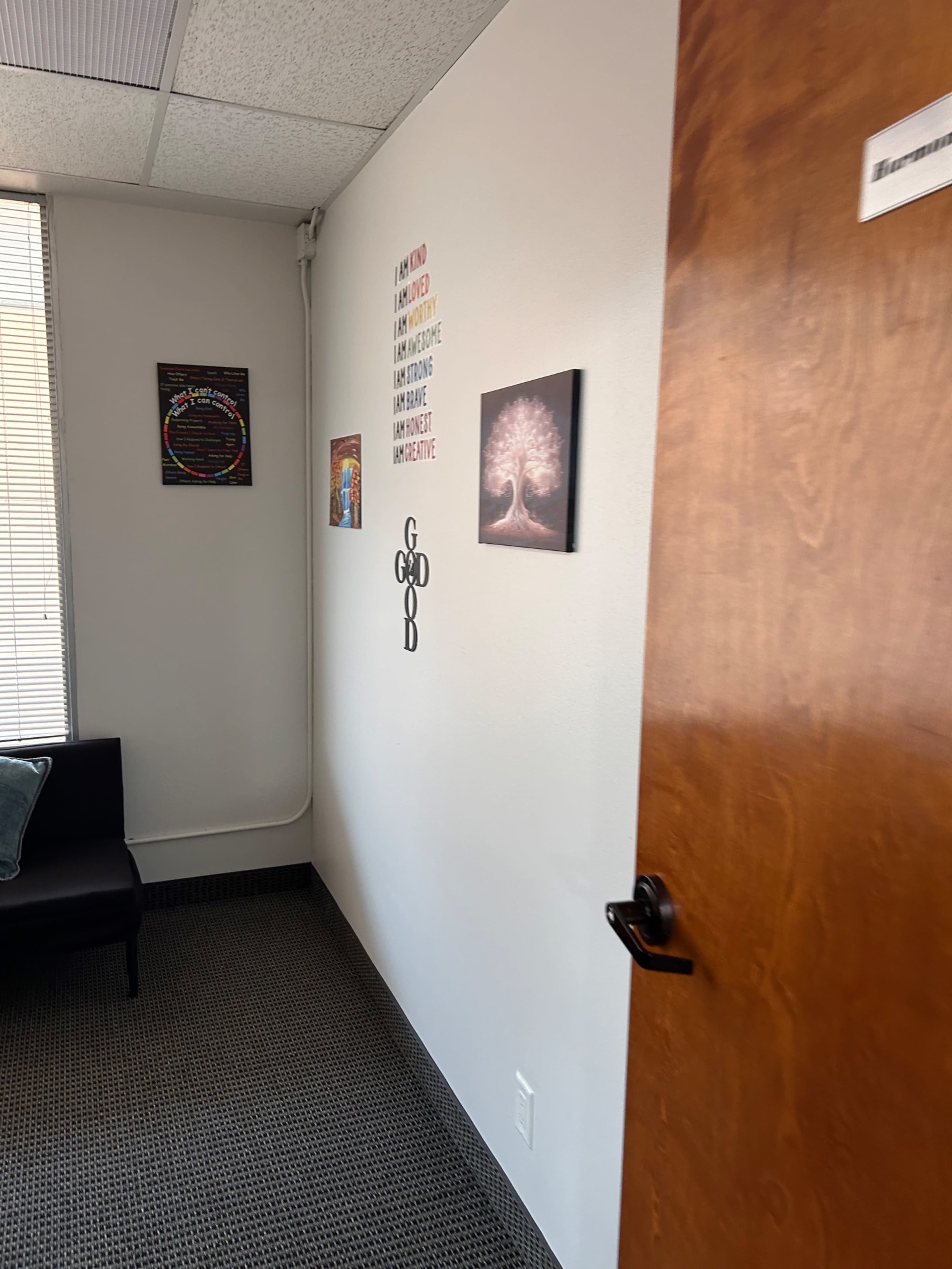 The image depicts a small office space with a dark couch against a wall, a closed wooden door, and framed artwork hanging on the walls.