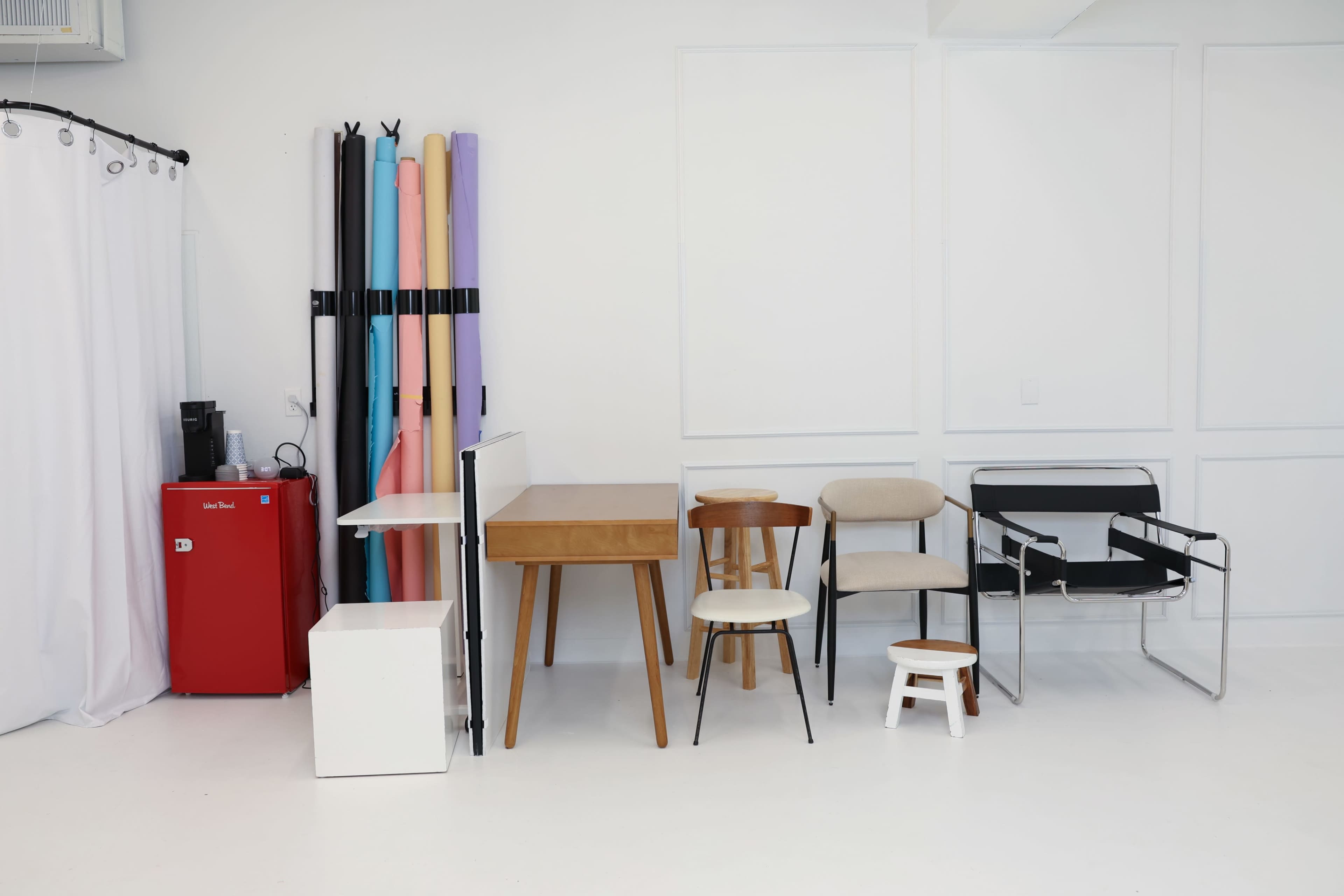 The image shows a minimalist studio setting with various chairs and a table arranged against a white wall, alongside a red refrigerator and colorful backdrop rolls.
