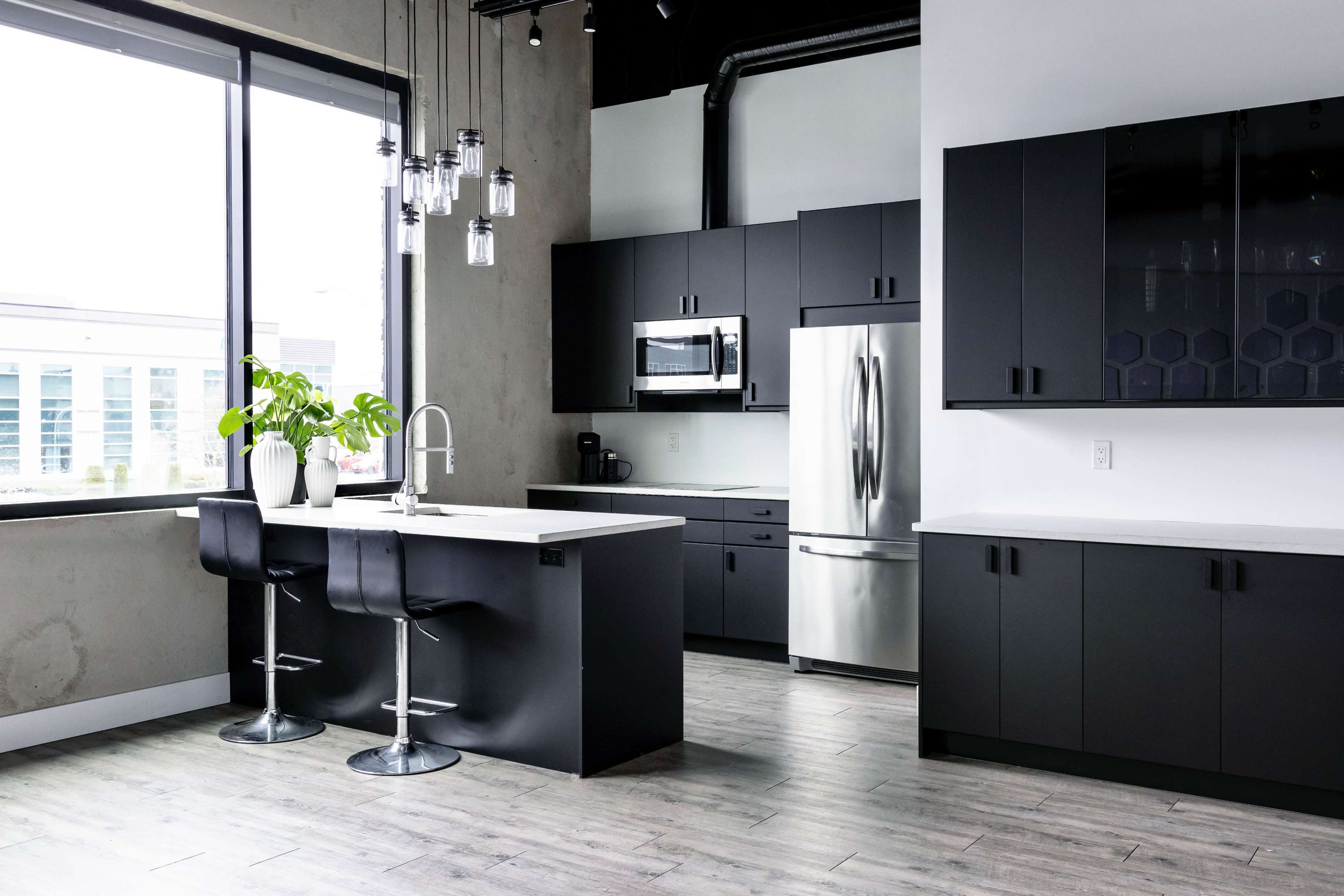 The image shows a modern kitchen with black cabinets, a stainless steel refrigerator, a sink, and pendant lighting over a central island with bar stools.