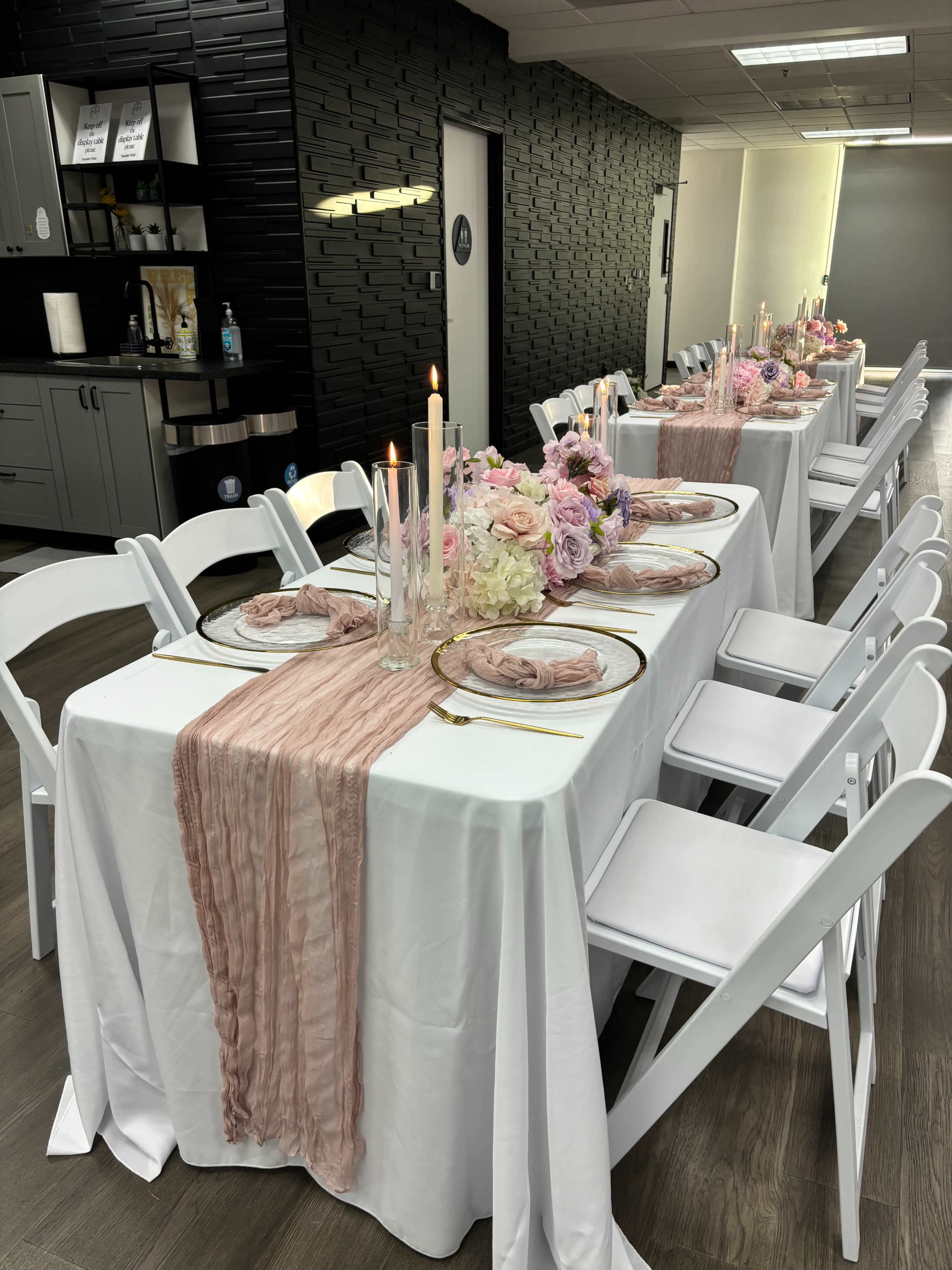 A long dining table is set with white linens, pink floral centerpieces, and candles, surrounded by white folding chairs in a well-lit room.