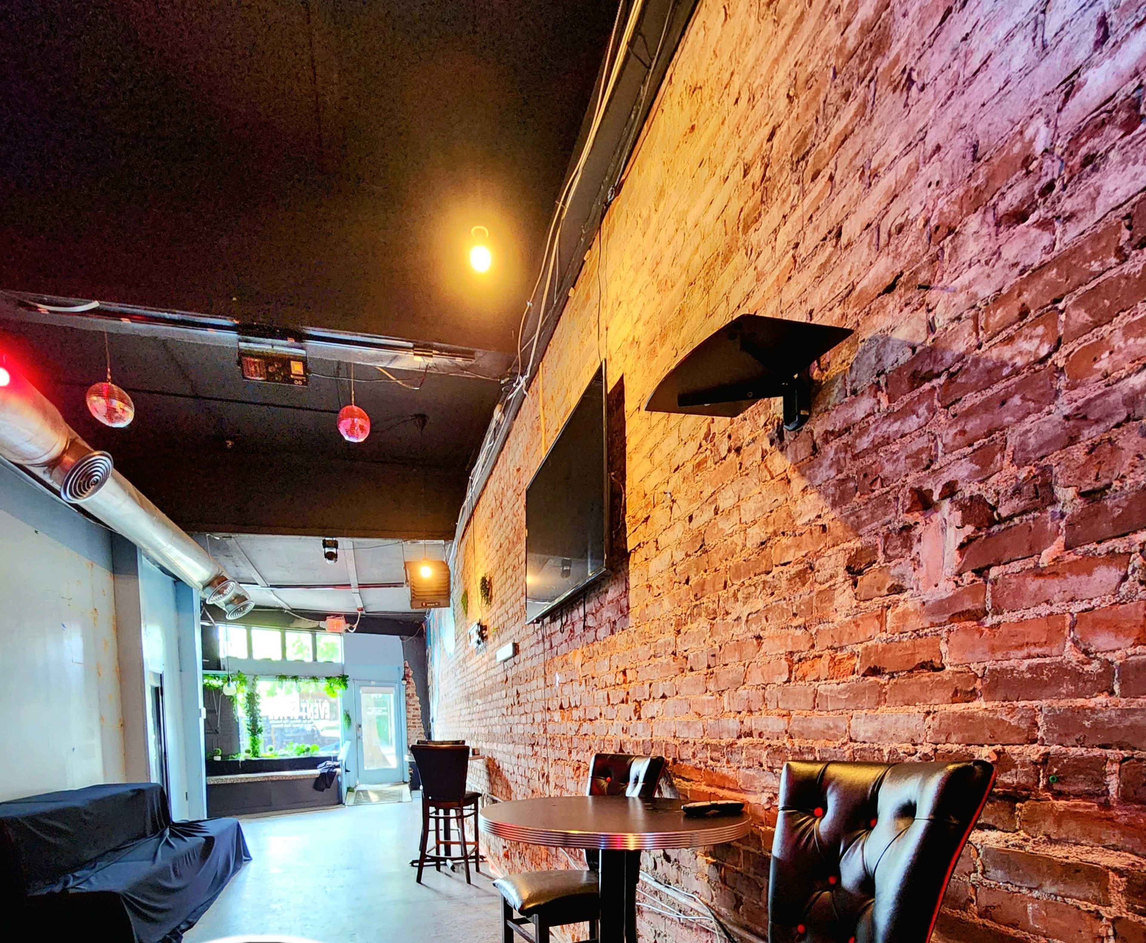 The image shows a dimly lit interior of a bar or lounge, featuring exposed brick walls, a flat-screen TV mounted on the wall, and a small table with a black leather chair.