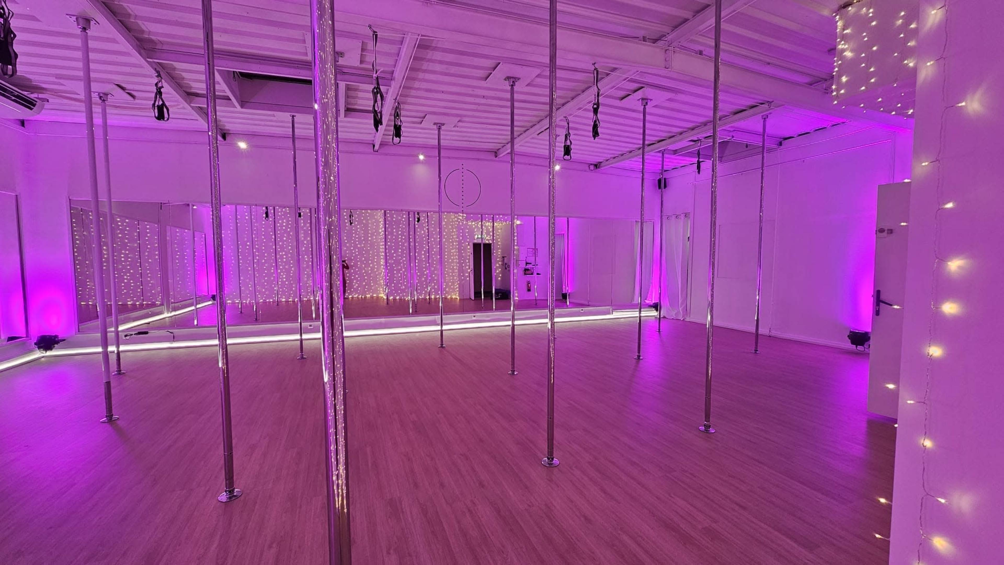A brightly lit dance studio featuring multiple silver poles and purple ambient lighting, with mirrored walls and wooden flooring.