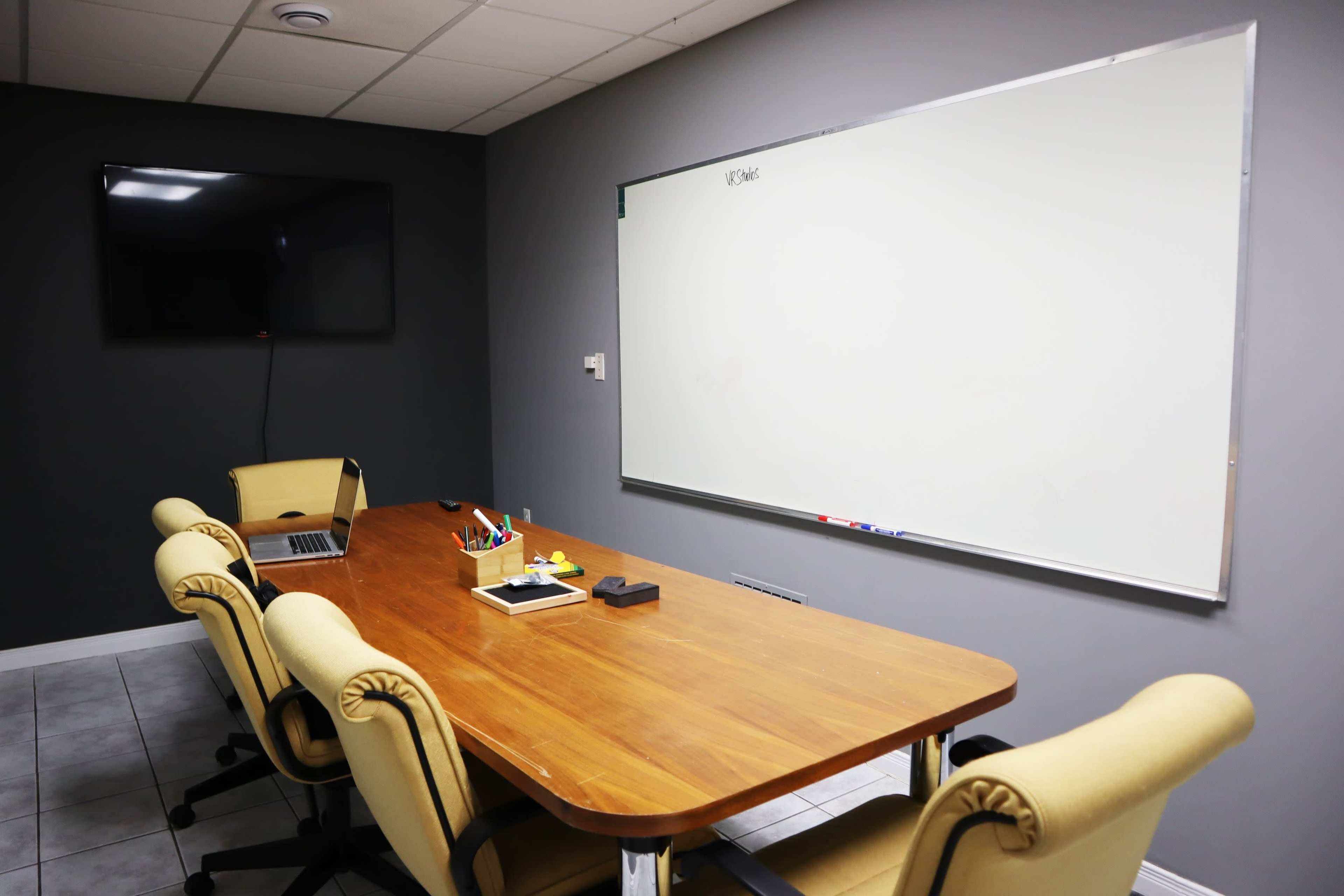 A conference room features a wooden table with six beige chairs, a laptop, and a whiteboard on the wall.