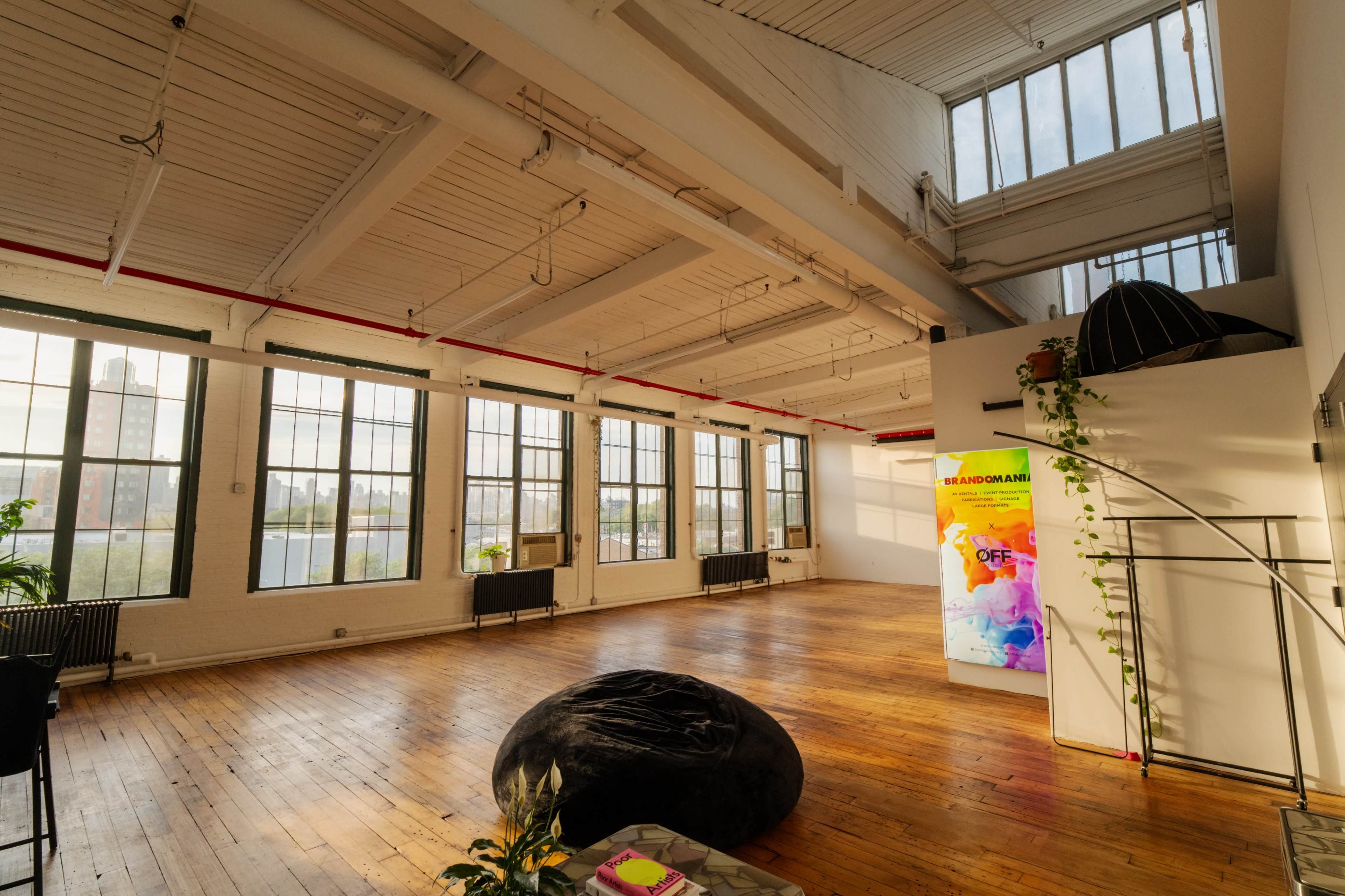 The image shows a spacious, airy room with large windows, wooden flooring, and minimal furniture, featuring a colorful wall display and a bean bag chair.