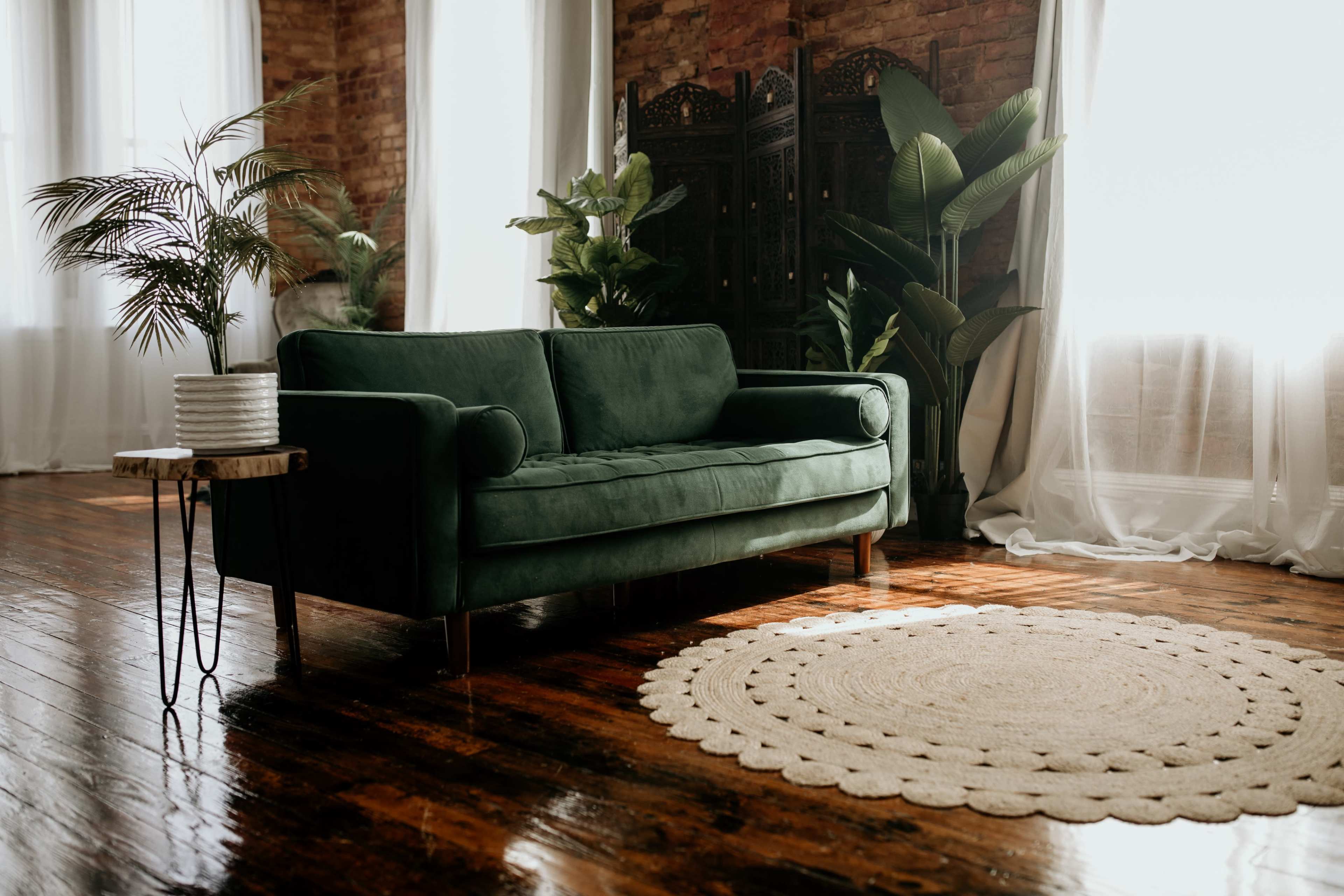 A green velvet sofa is positioned near a round woven rug in a room with large windows and potted plants.