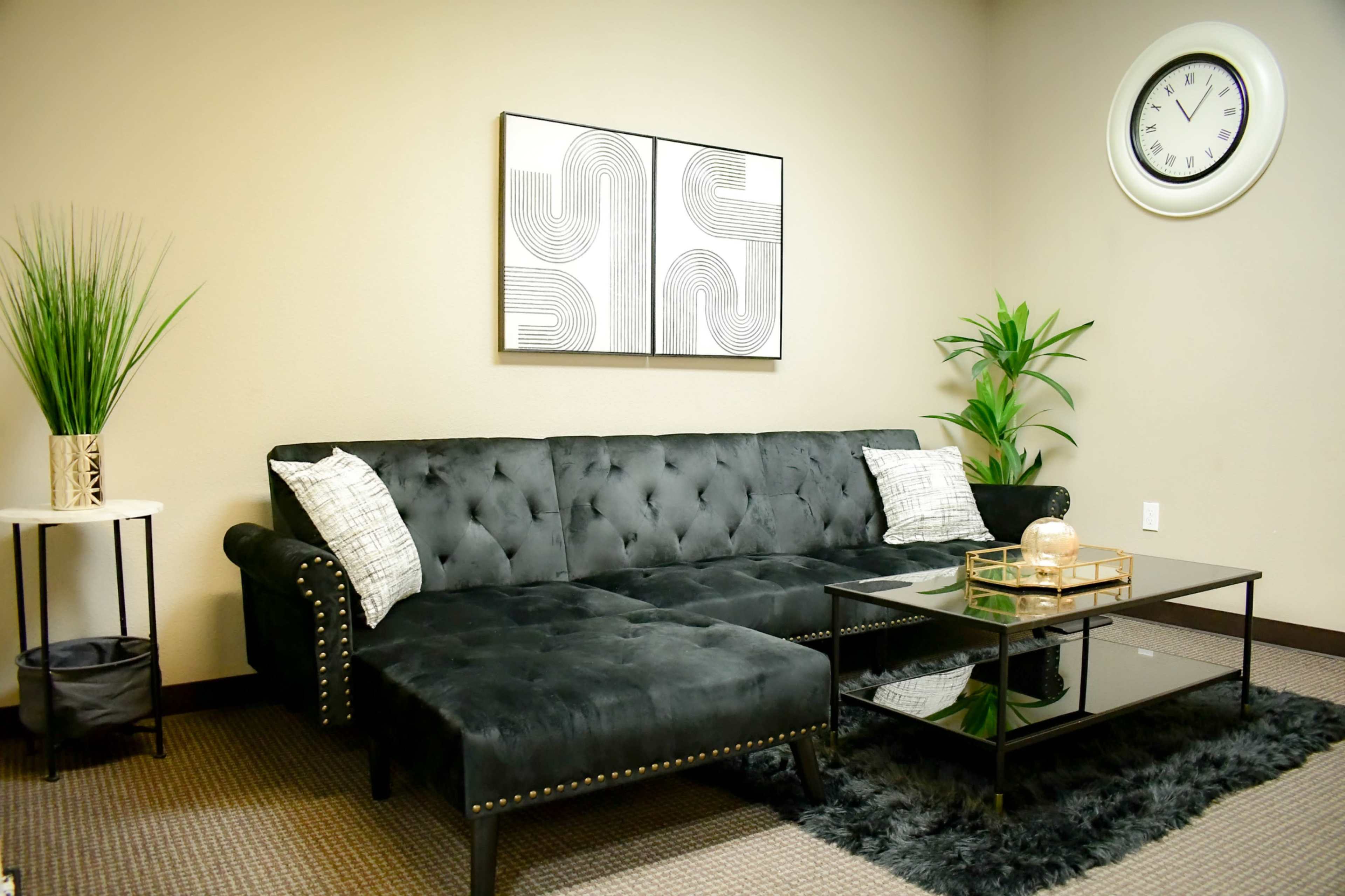 A modern living room featuring a black tufted sectional sofa, a glass coffee table, and decorative plants against a neutral wall.