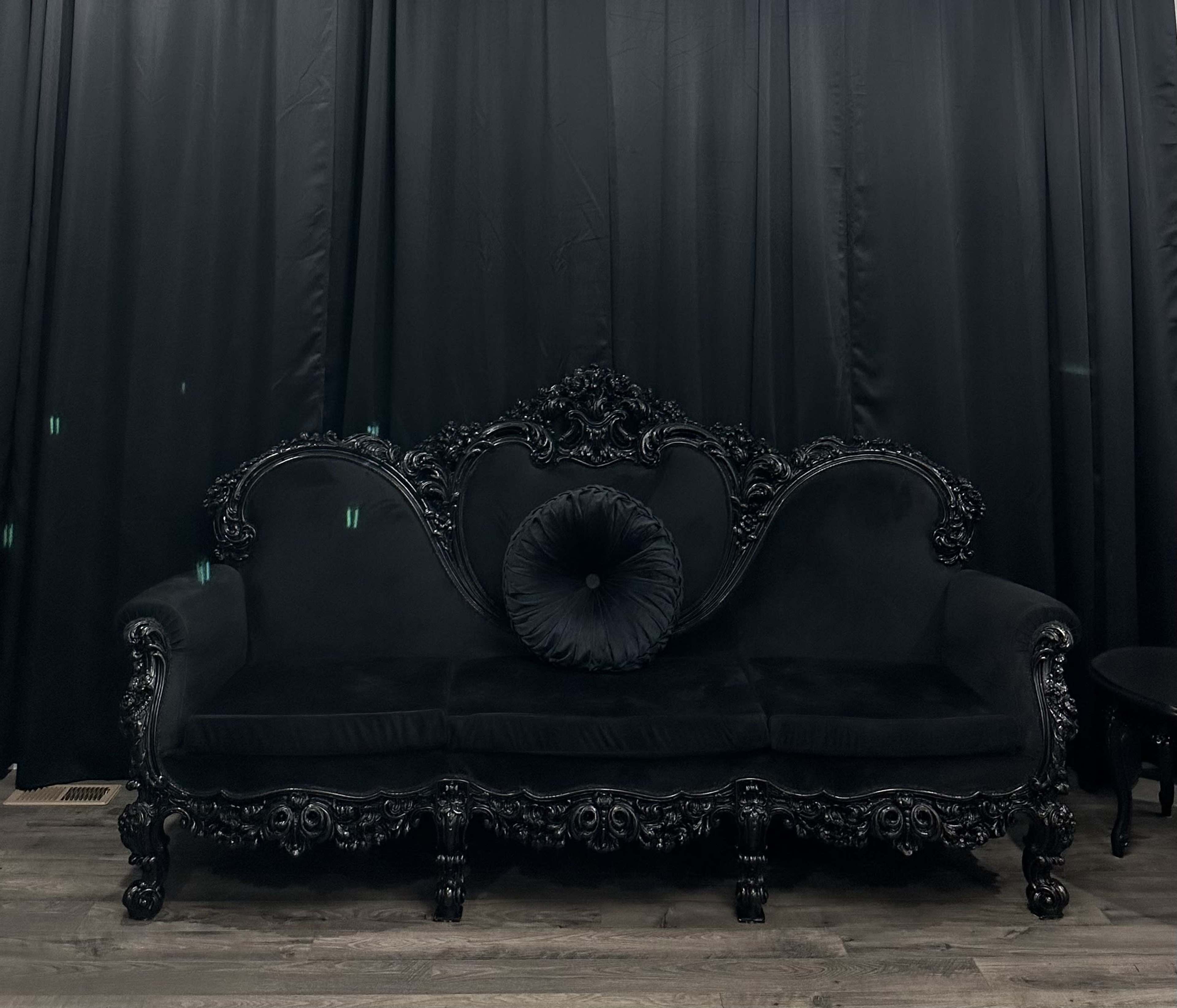 An ornate black velvet sofa with a decorative backrest and a central round cushion, set against a black curtain backdrop.