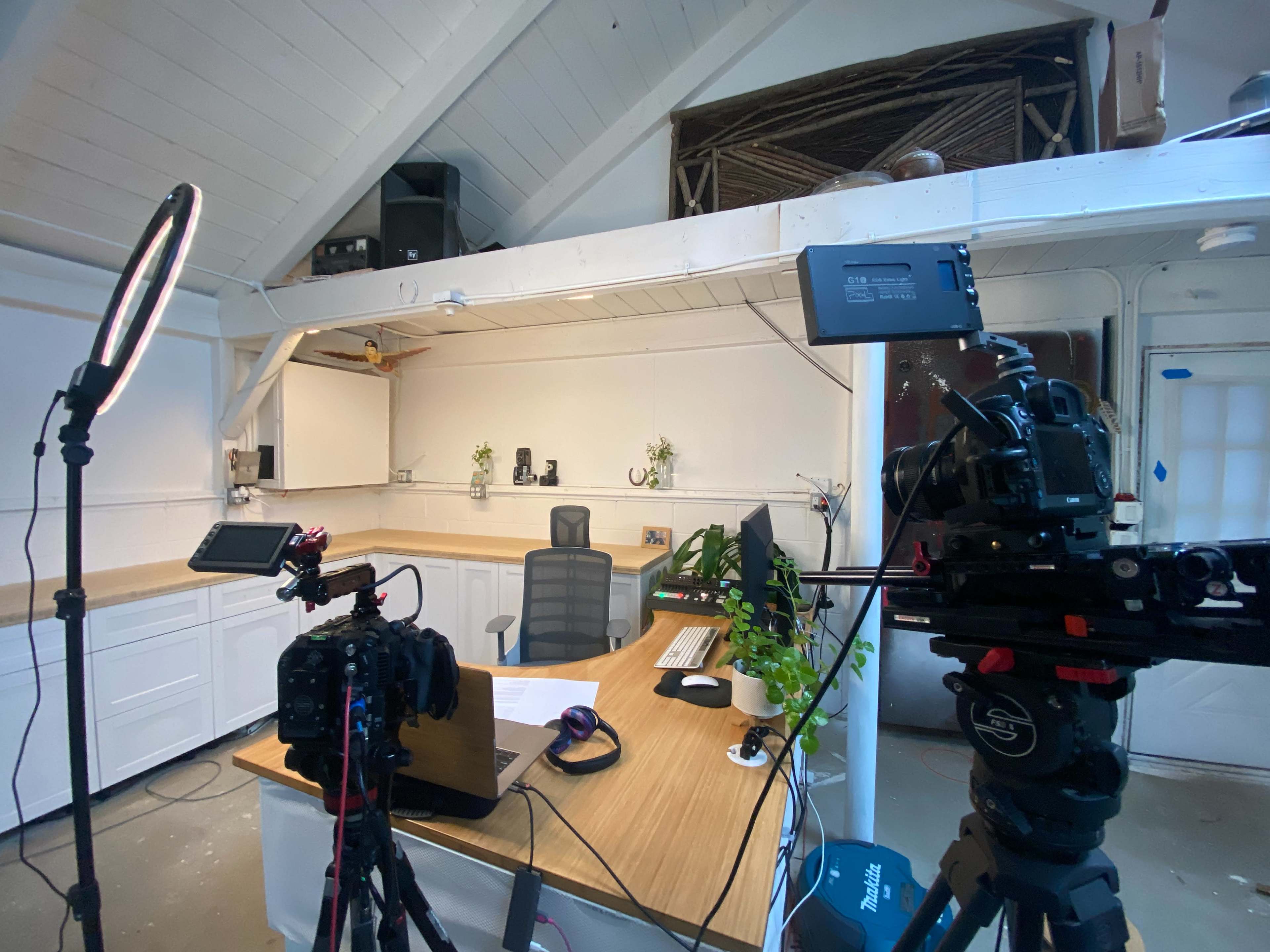 A modern office space is set up for filming, featuring a wooden desk with a laptop, several cameras on tripods, and lighting equipment arranged around the area.
