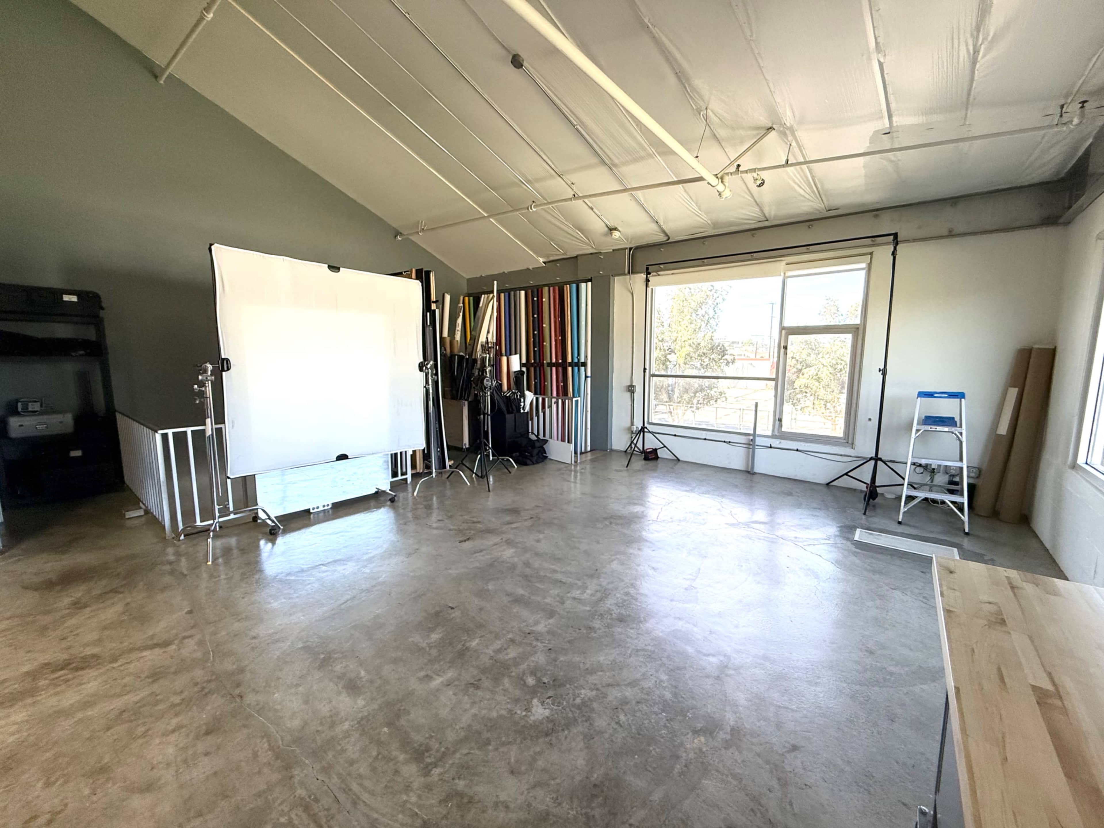 The image shows a spacious, unfinished studio with a concrete floor, backdrop stands, and a large window letting in natural light.