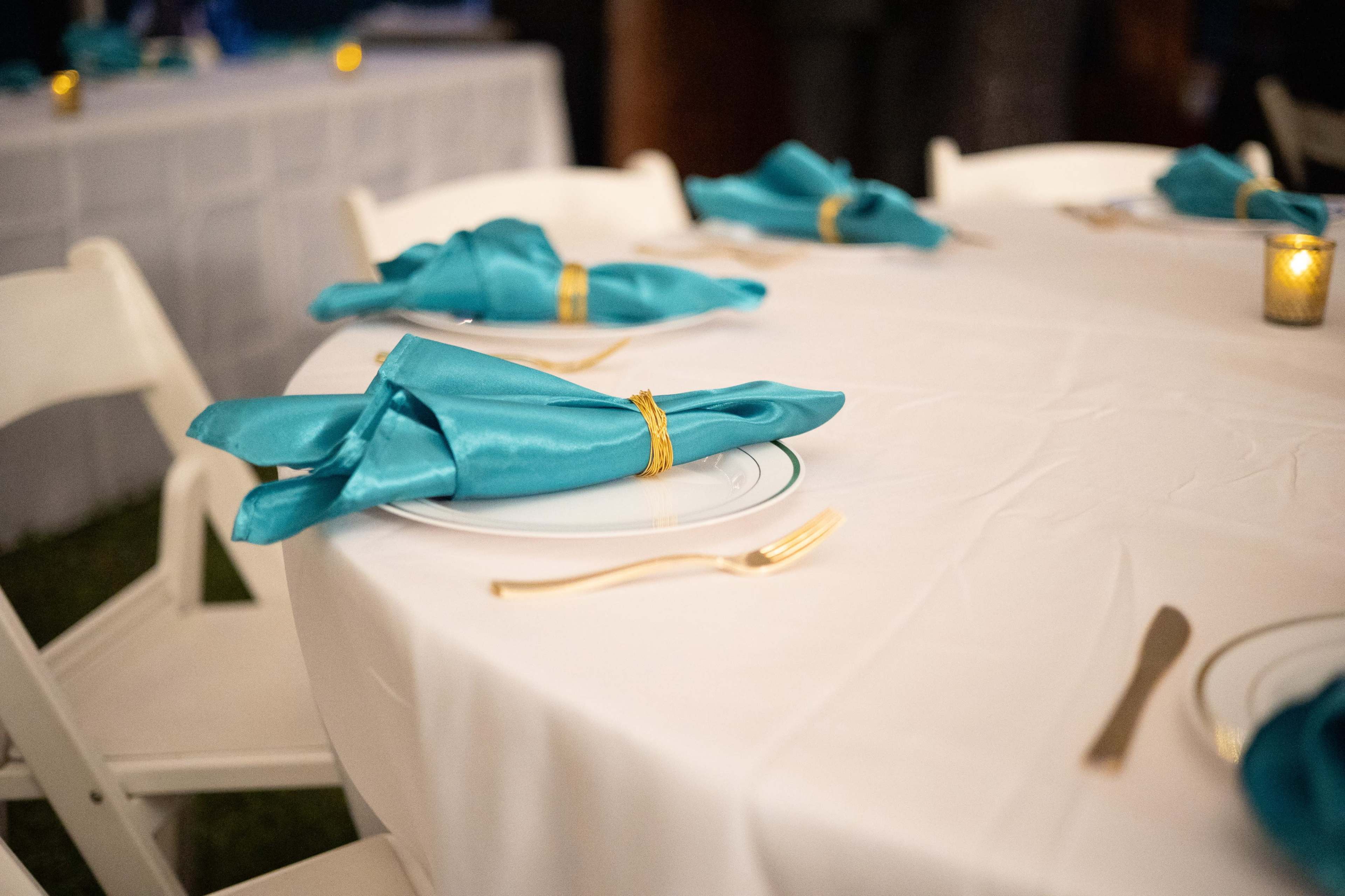A round table is set with white tablecloths, turquoise napkins neatly folded and placed on plates, accompanied by golden flatware.