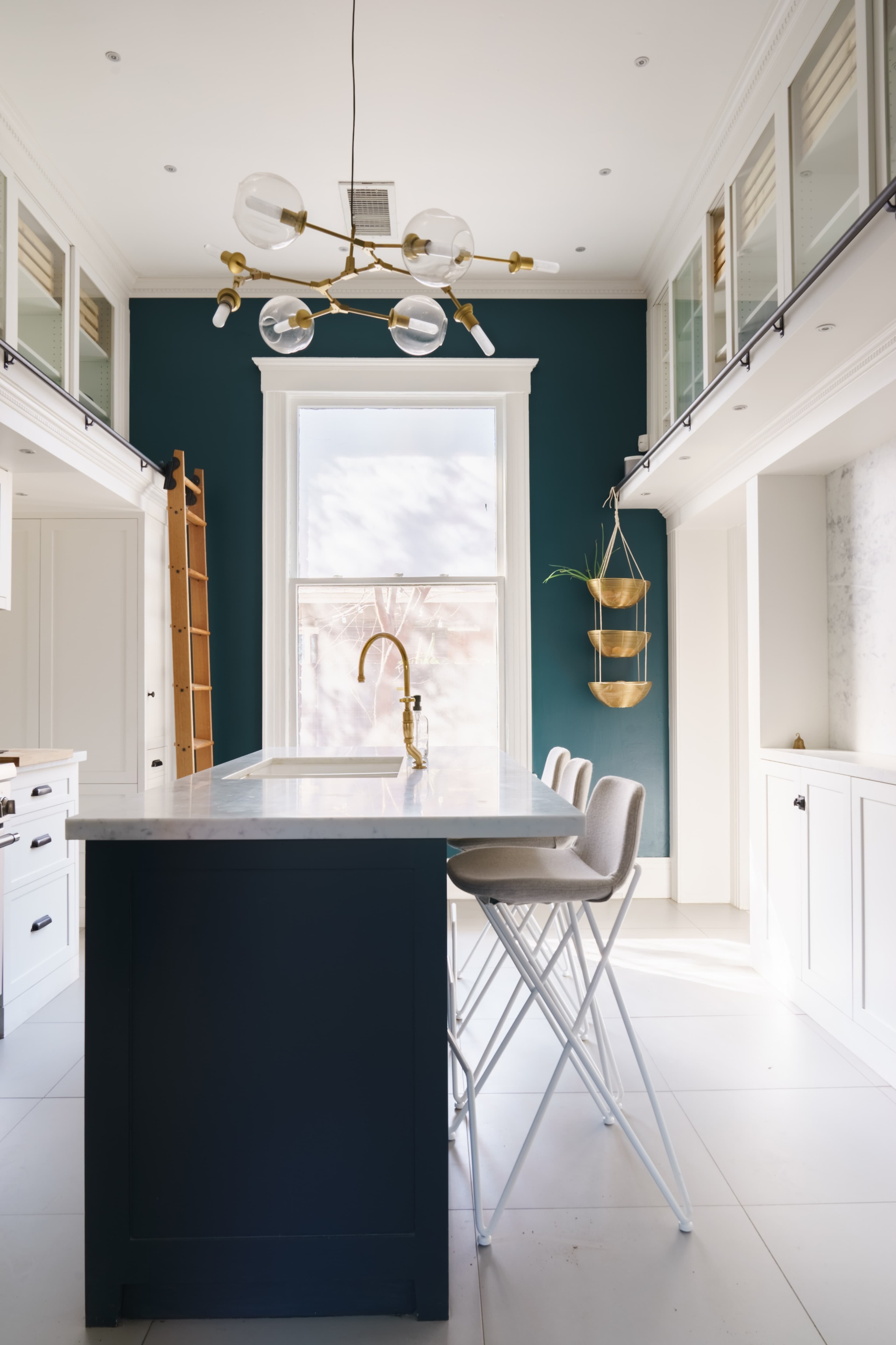 A modern kitchen featuring a dark blue island with a marble countertop, white cabinetry, and a large window with brass fixtures.