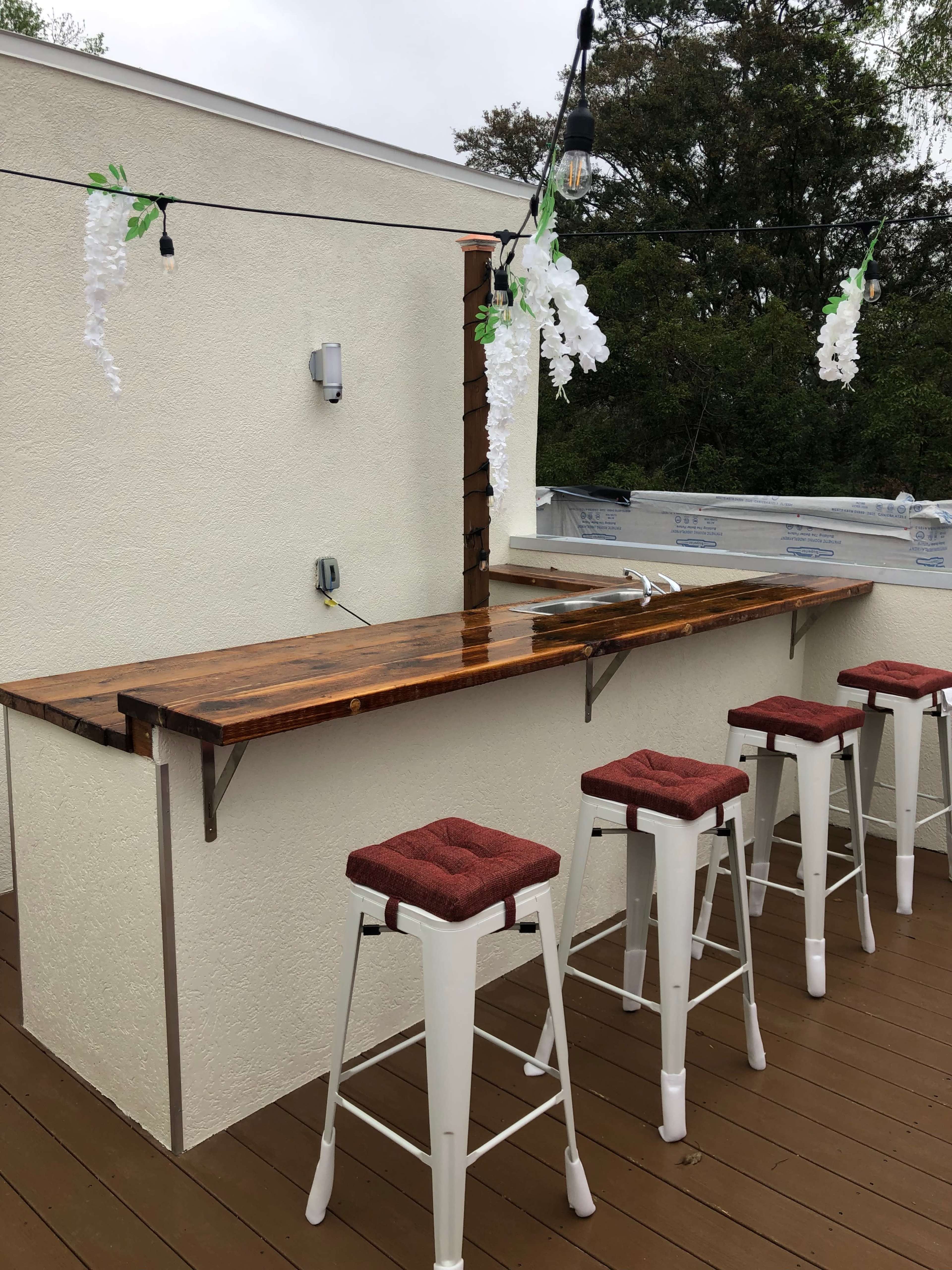 A wooden bar with red cushioned stools is set up outdoors under string lights and decorative hanging flowers.