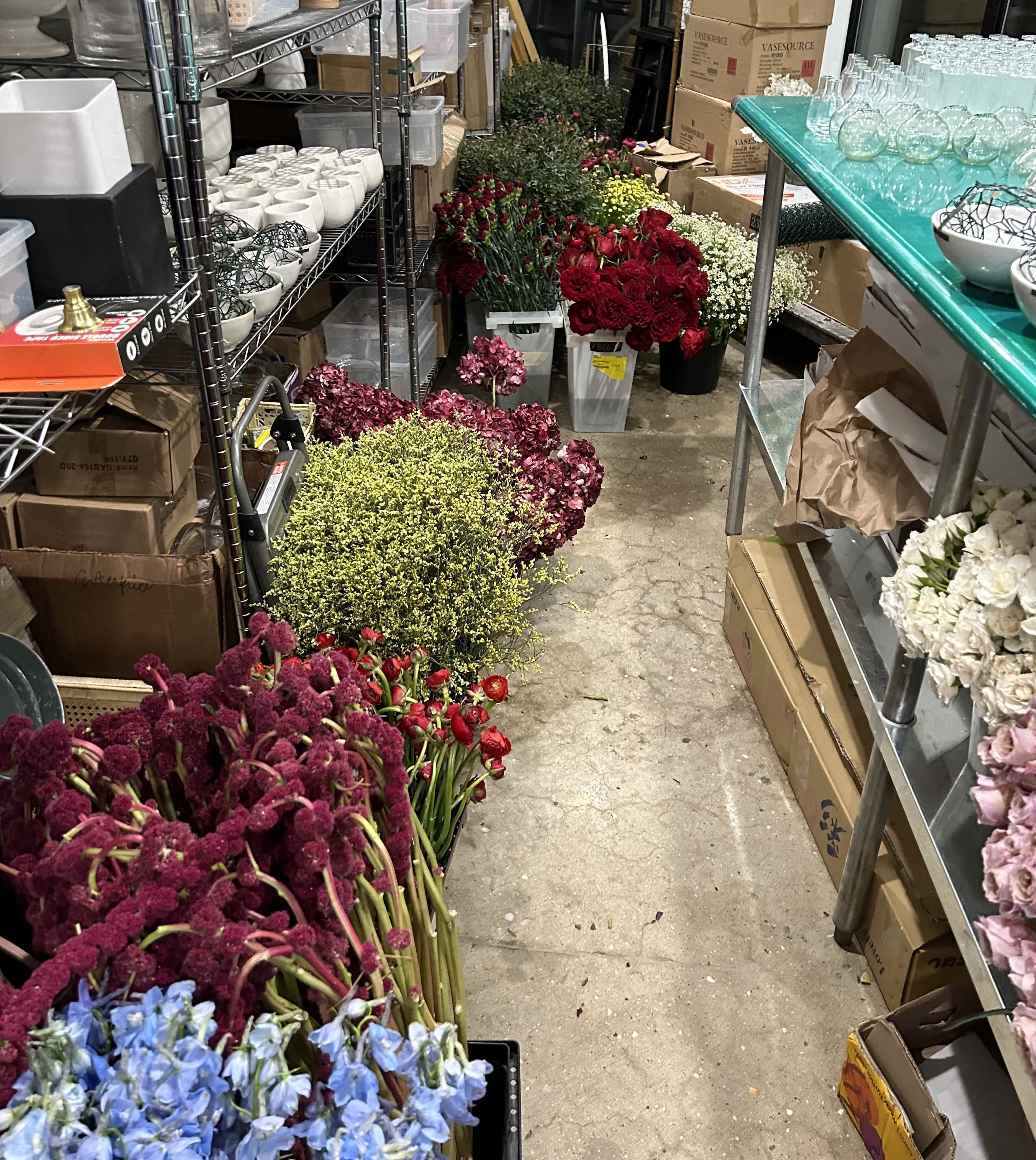 The image shows a storage area organized with shelves stacked with various boxes, and an assortment of flowers in different colors and arrangements placed along the floor.