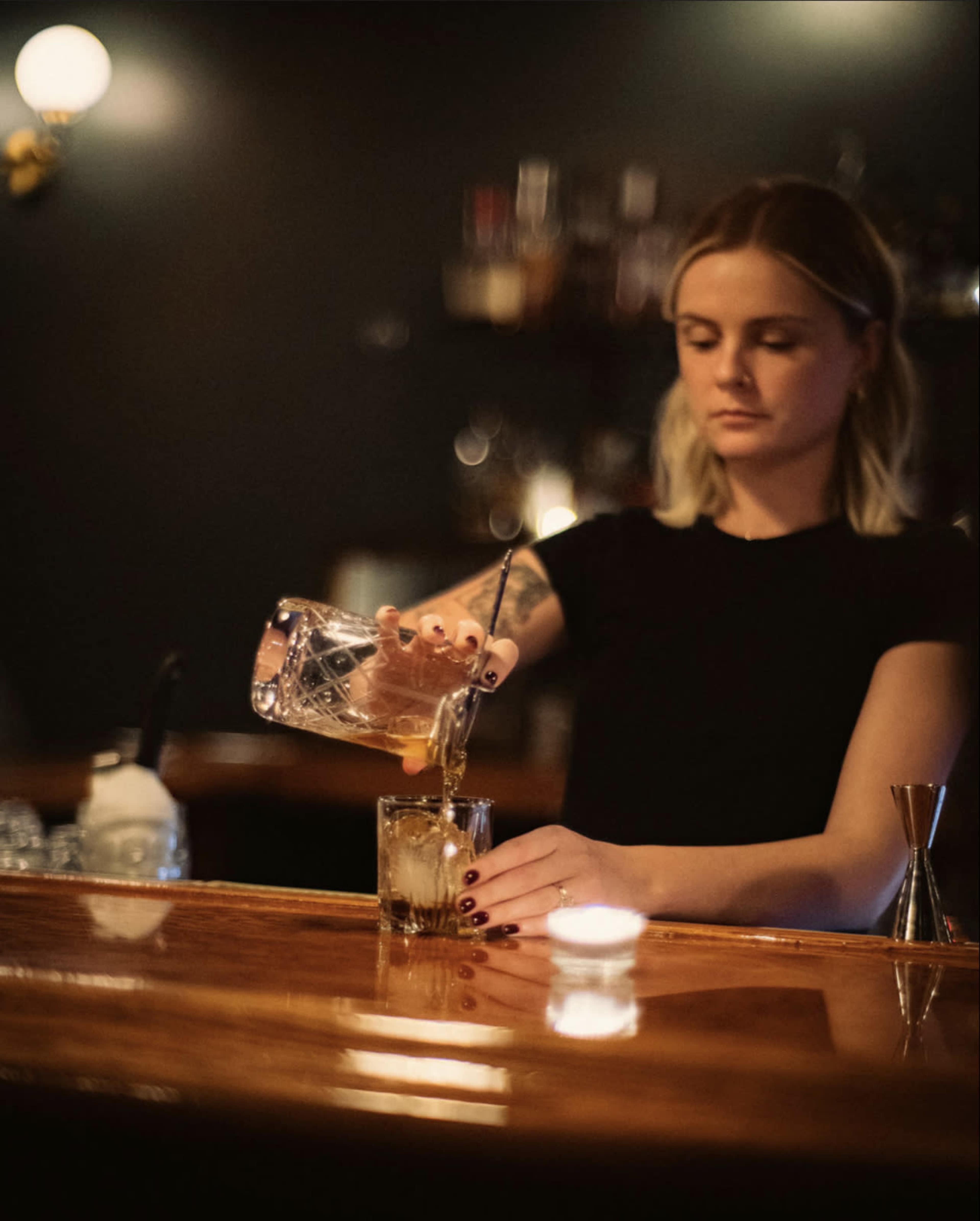 A bartender pours a drink into a glass filled with ice at a dimly lit bar.