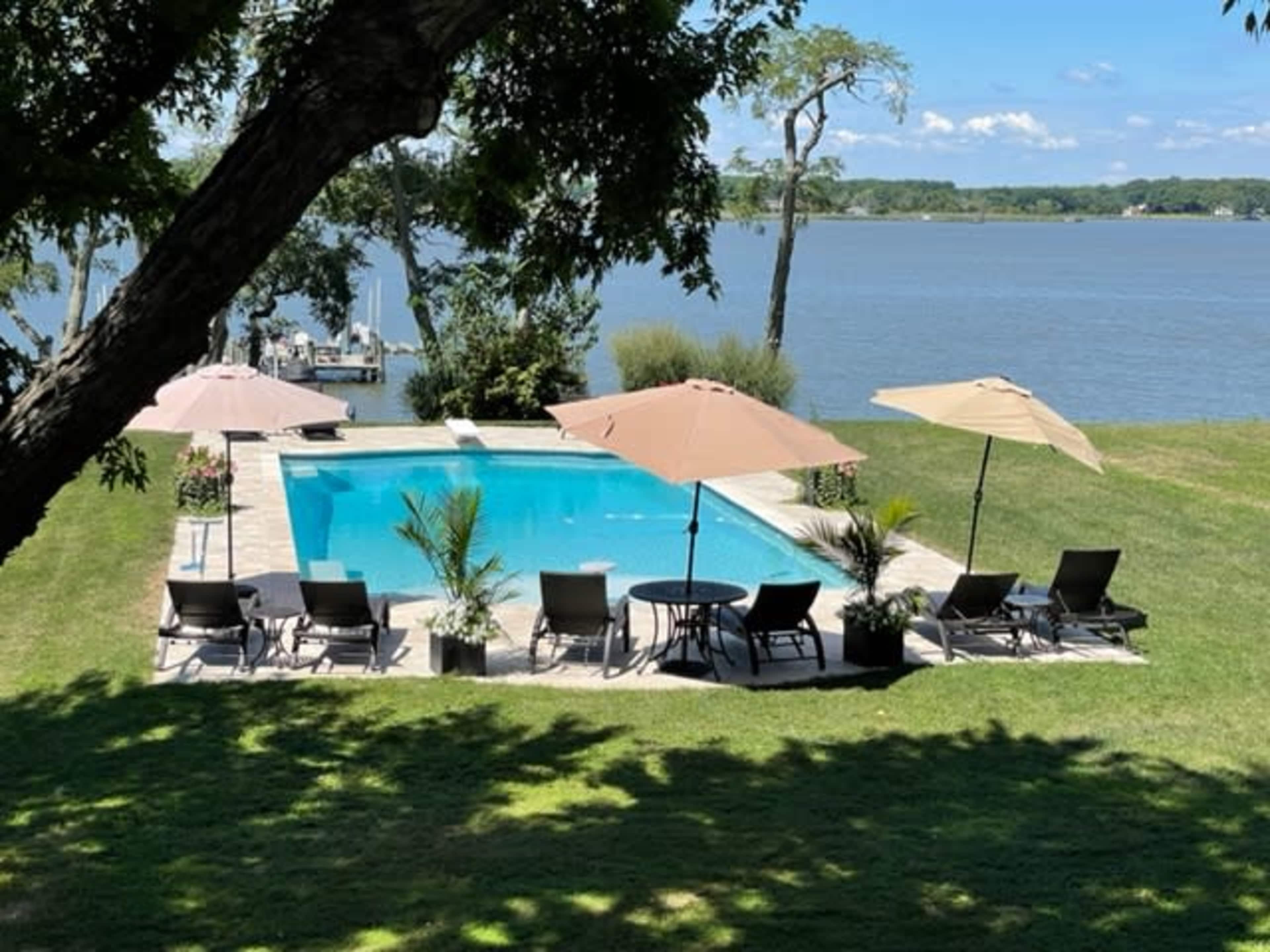 A rectangular swimming pool surrounded by lounge chairs and umbrellas, with a view of the water and greenery in the background.