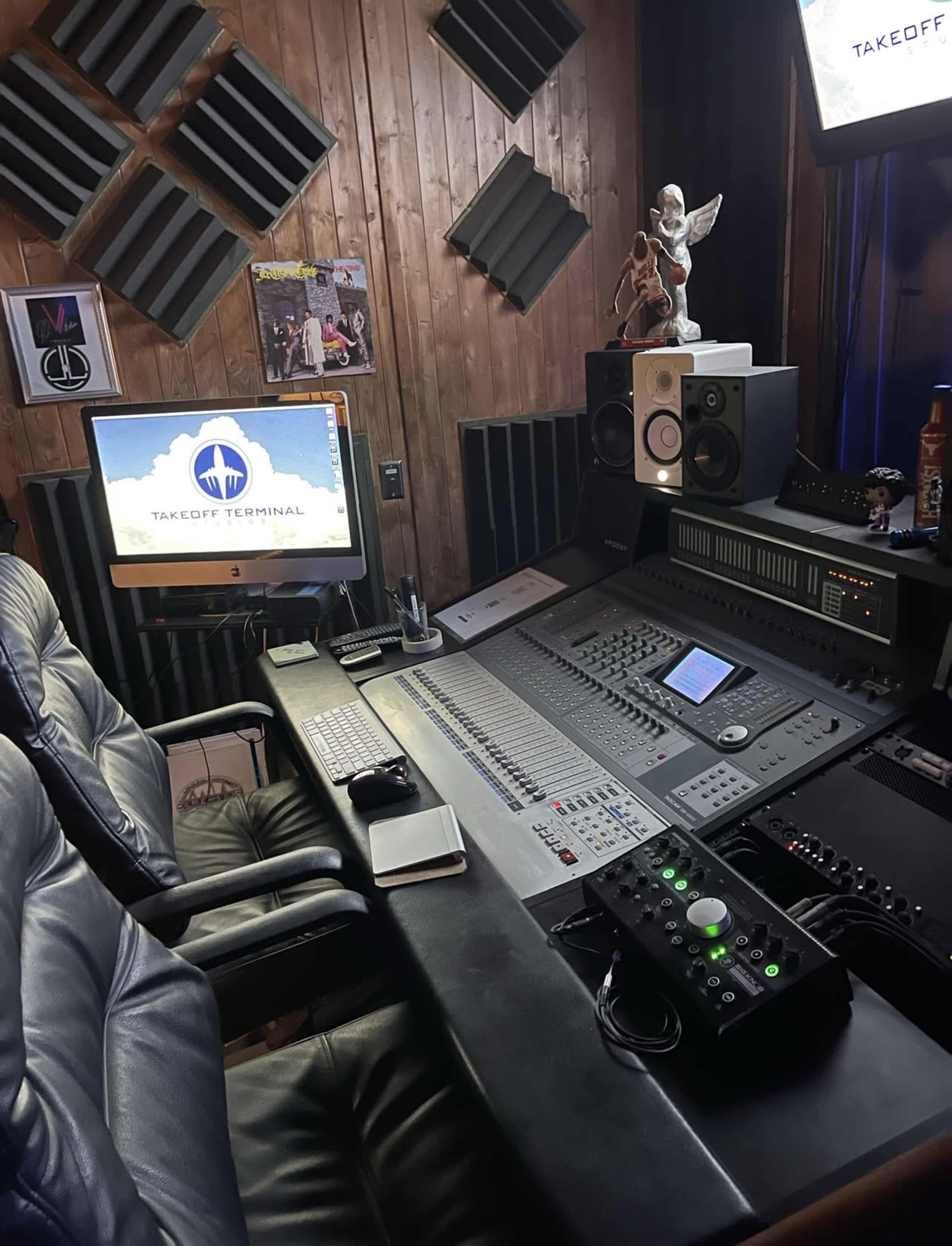 A music studio control desk with a computer, sound mixing equipment, and a black leather chair in front of a wooden wall.