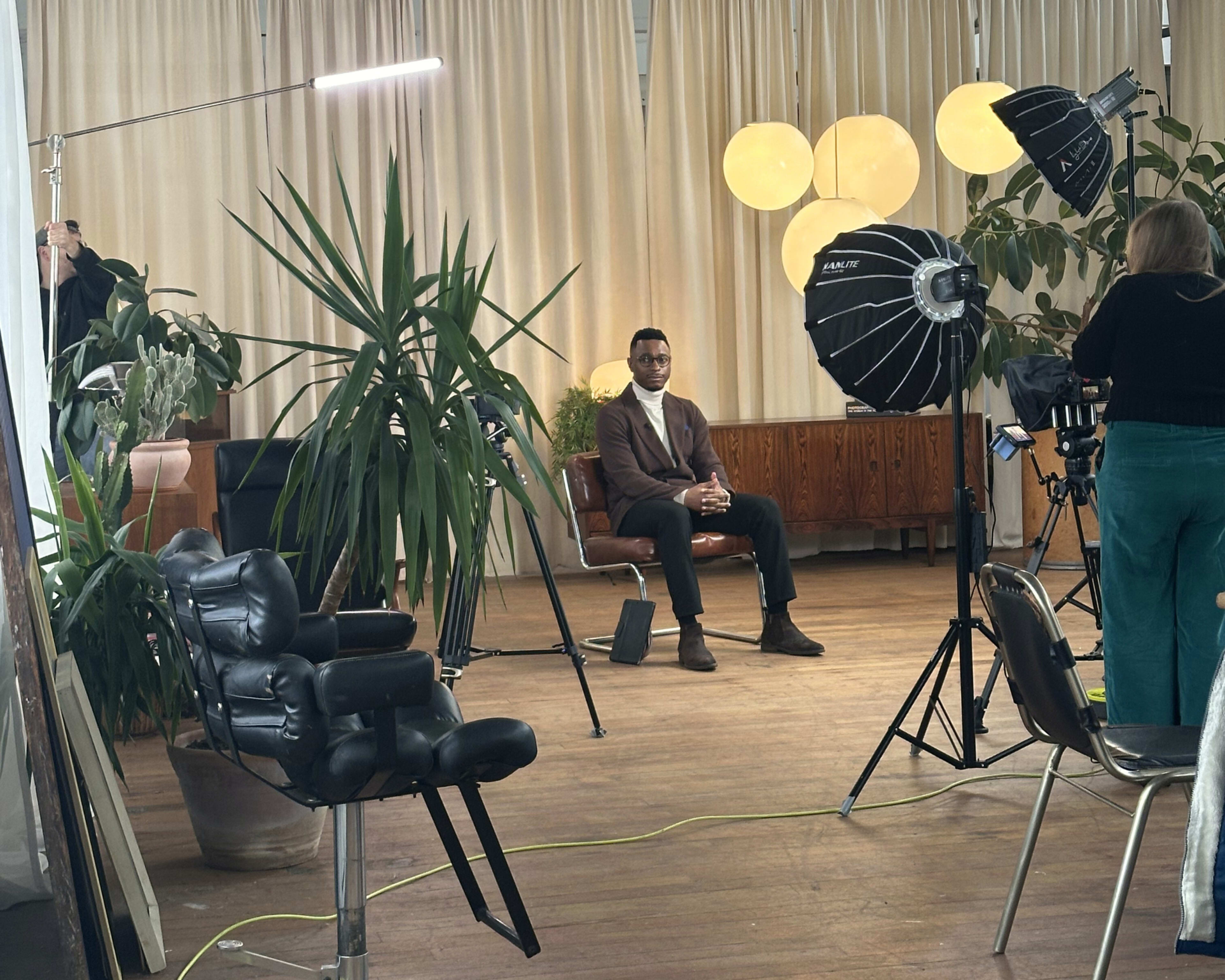 A man sits on a chair in front of large studio lights, surrounded by plants and a backdrop of curtains.