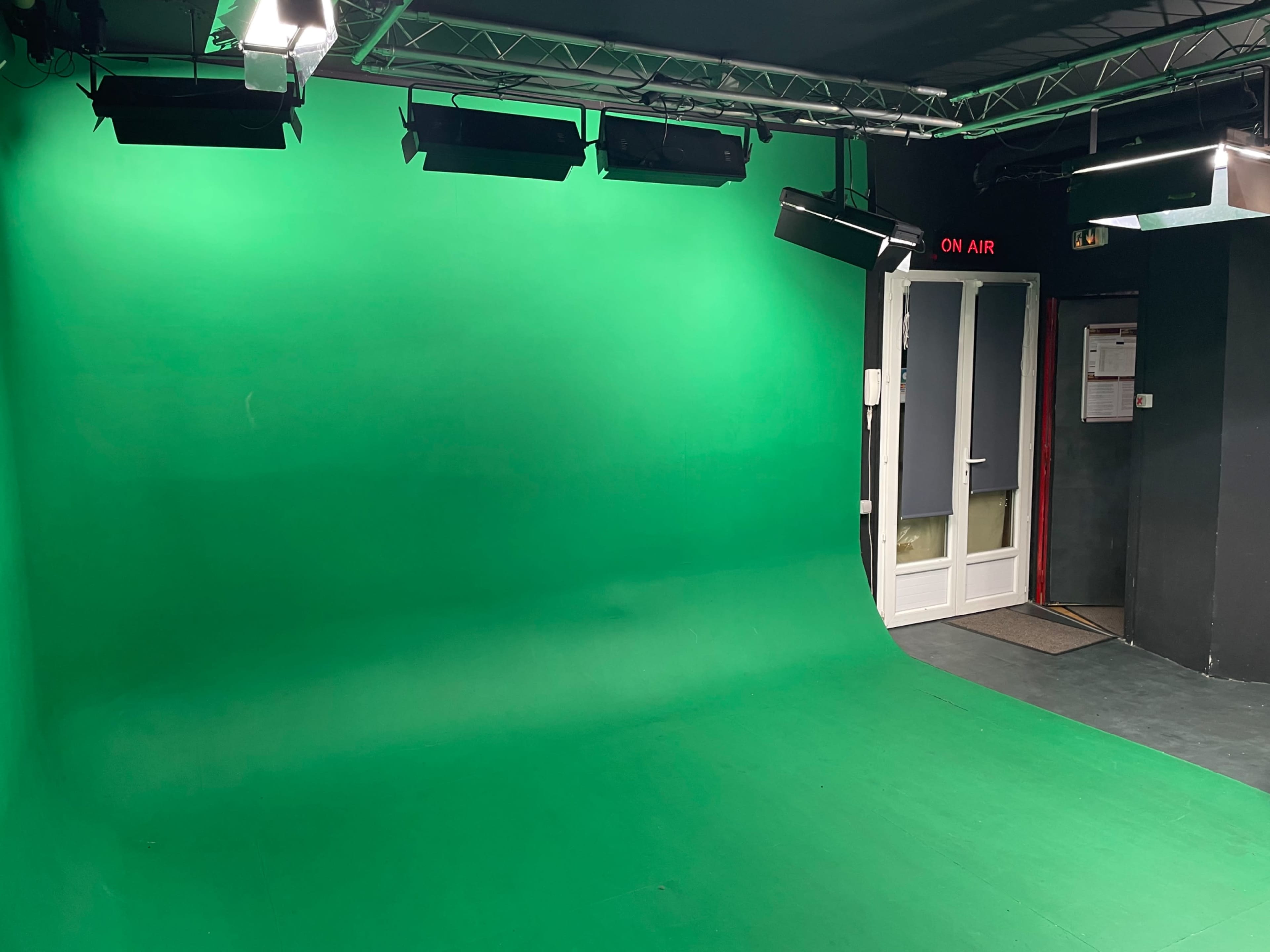The image shows a green screen studio with a seamless green backdrop, overhead lights, and a door labeled "ON AIR."