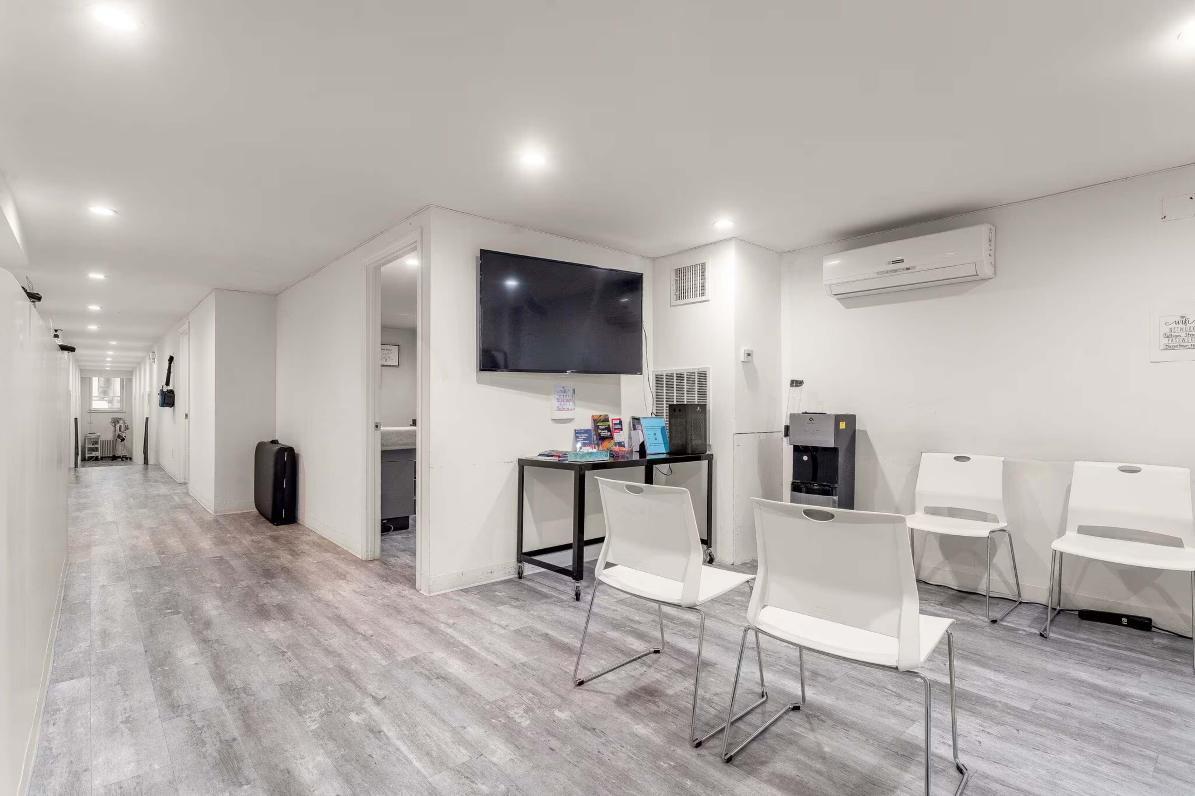 The image shows a modern, brightly lit waiting area with white chairs, a wall-mounted television, and a small table displaying brochures.