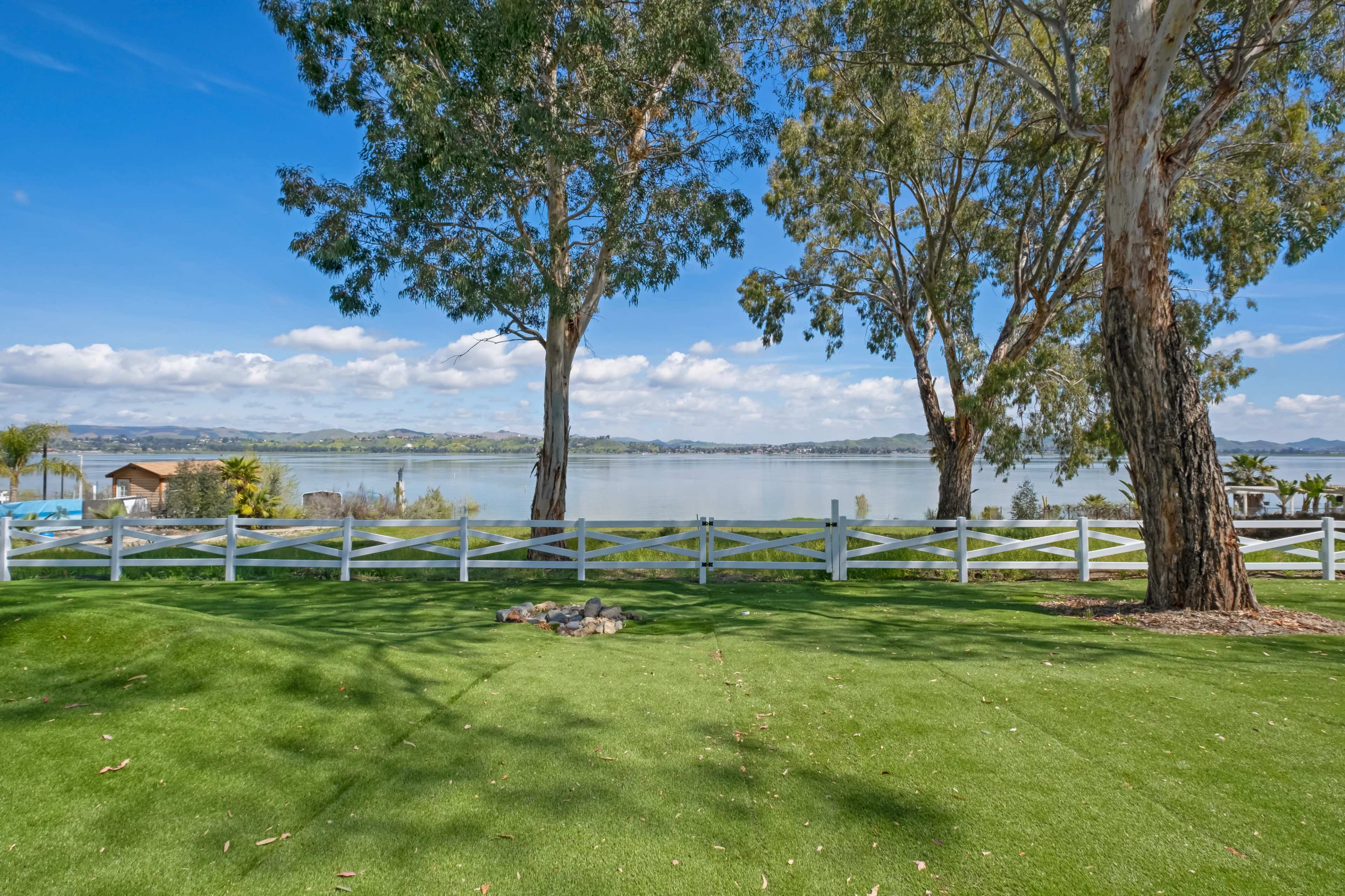 A tranquil lakeside scene with a green lawn, white fences, and trees framing the view of the water and distant hills.