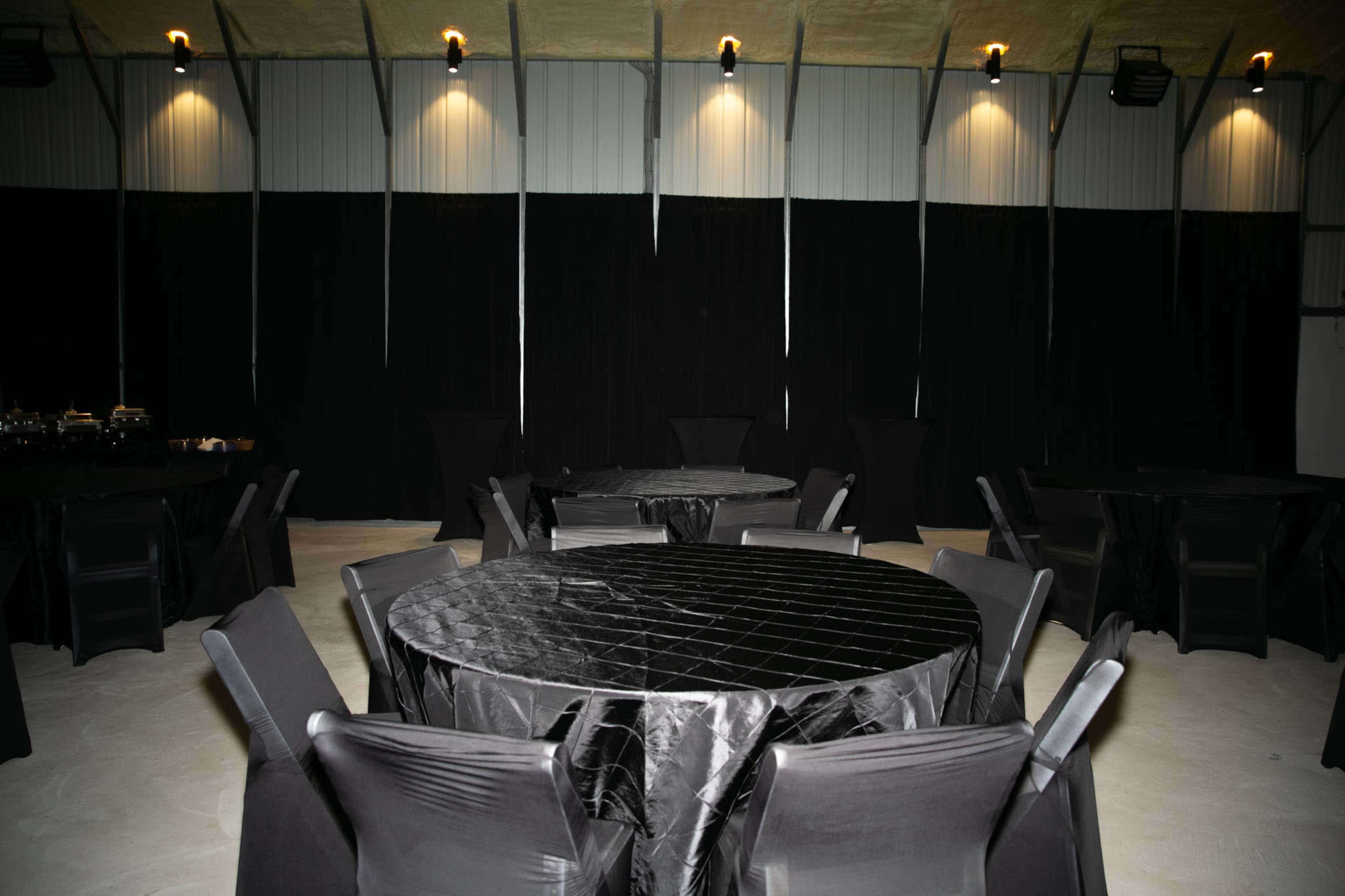 The image shows a spacious venue with several round tables covered in black tablecloths, surrounded by black folding chairs against a backdrop of black curtains.