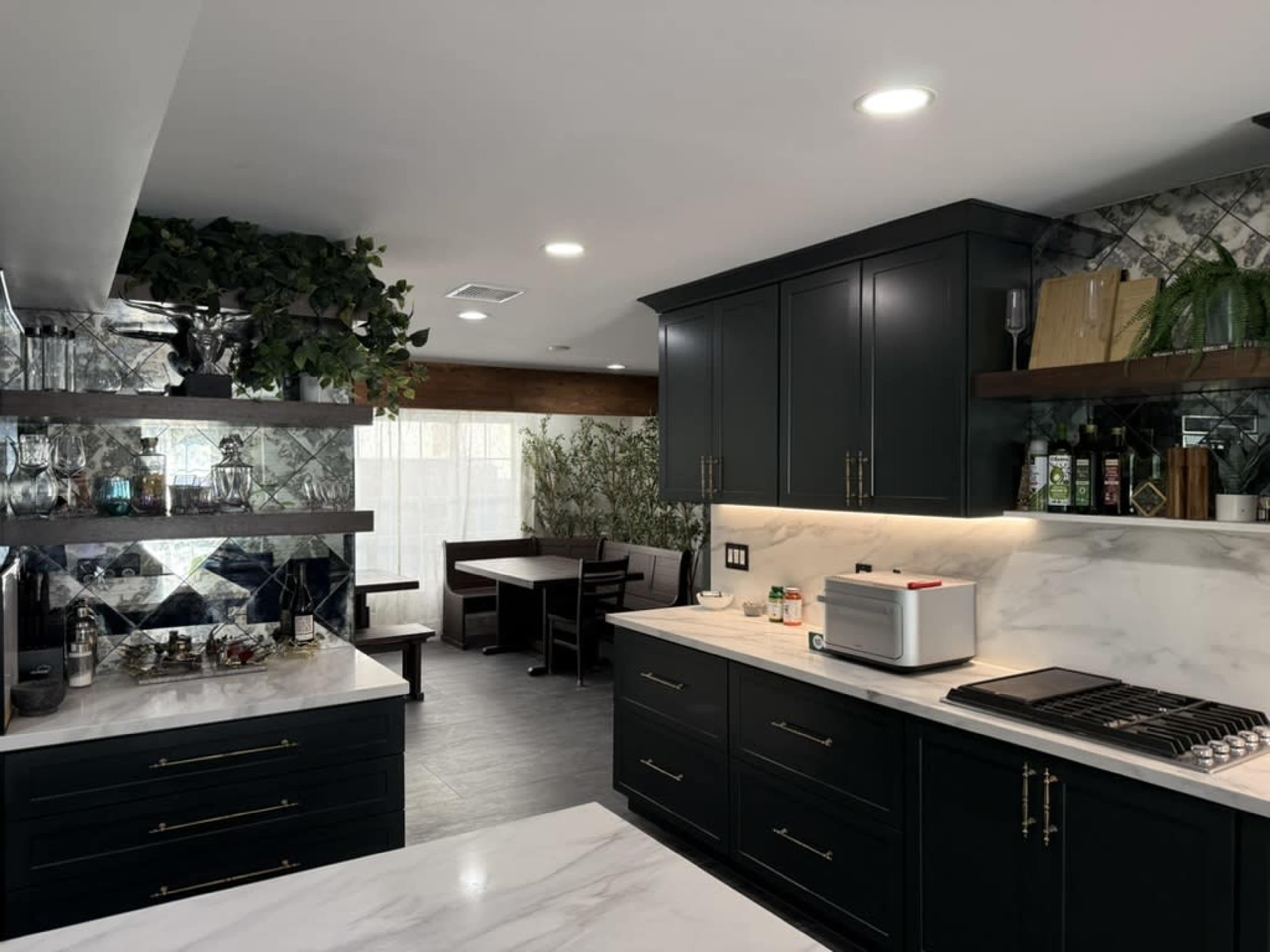 The image shows a modern kitchen with dark cabinets, marble countertops, and an adjoining dining area featuring a wooden table and benches.
