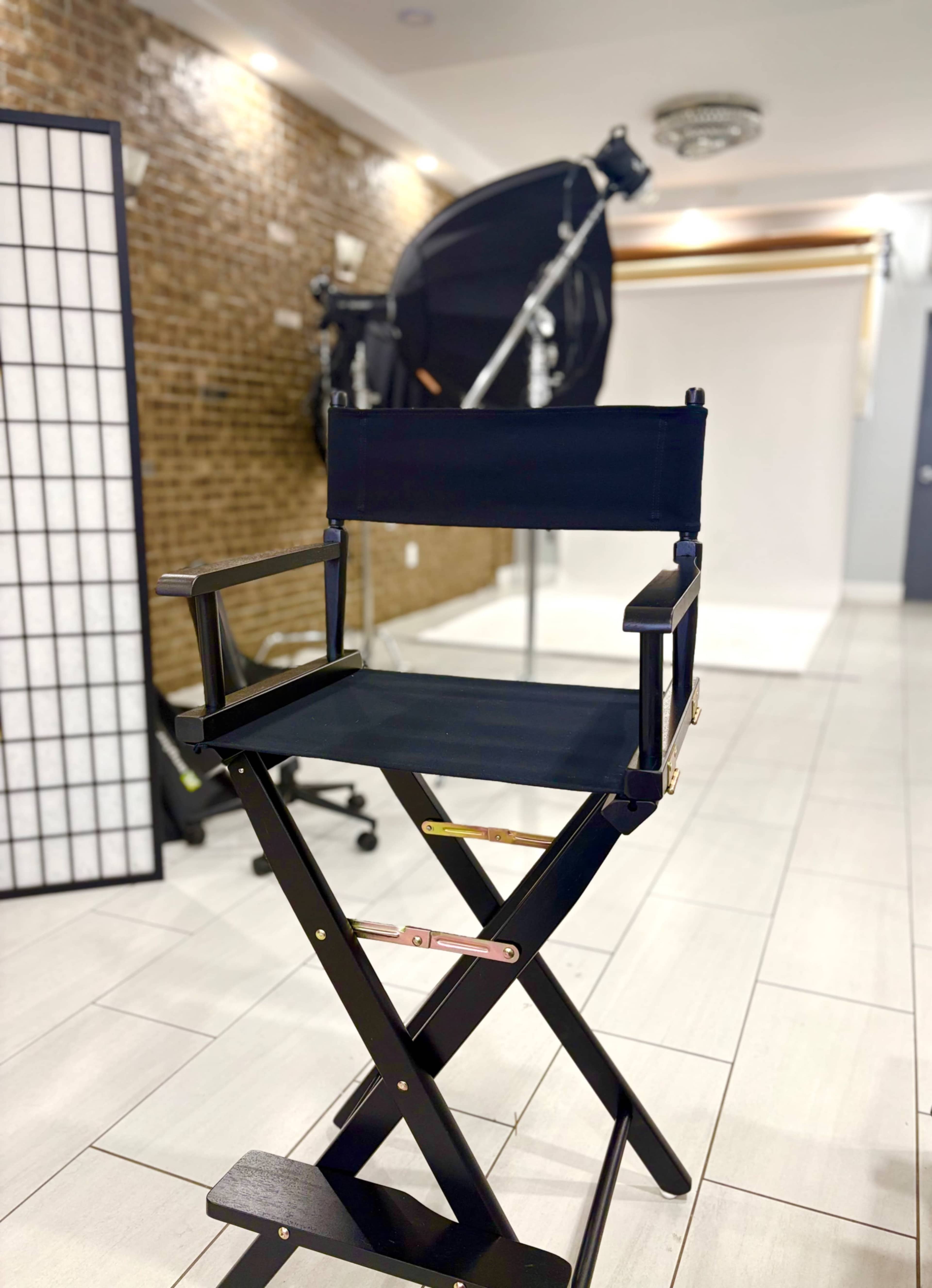 A black director's chair is positioned in a studio space featuring a light setup and a backdrop.