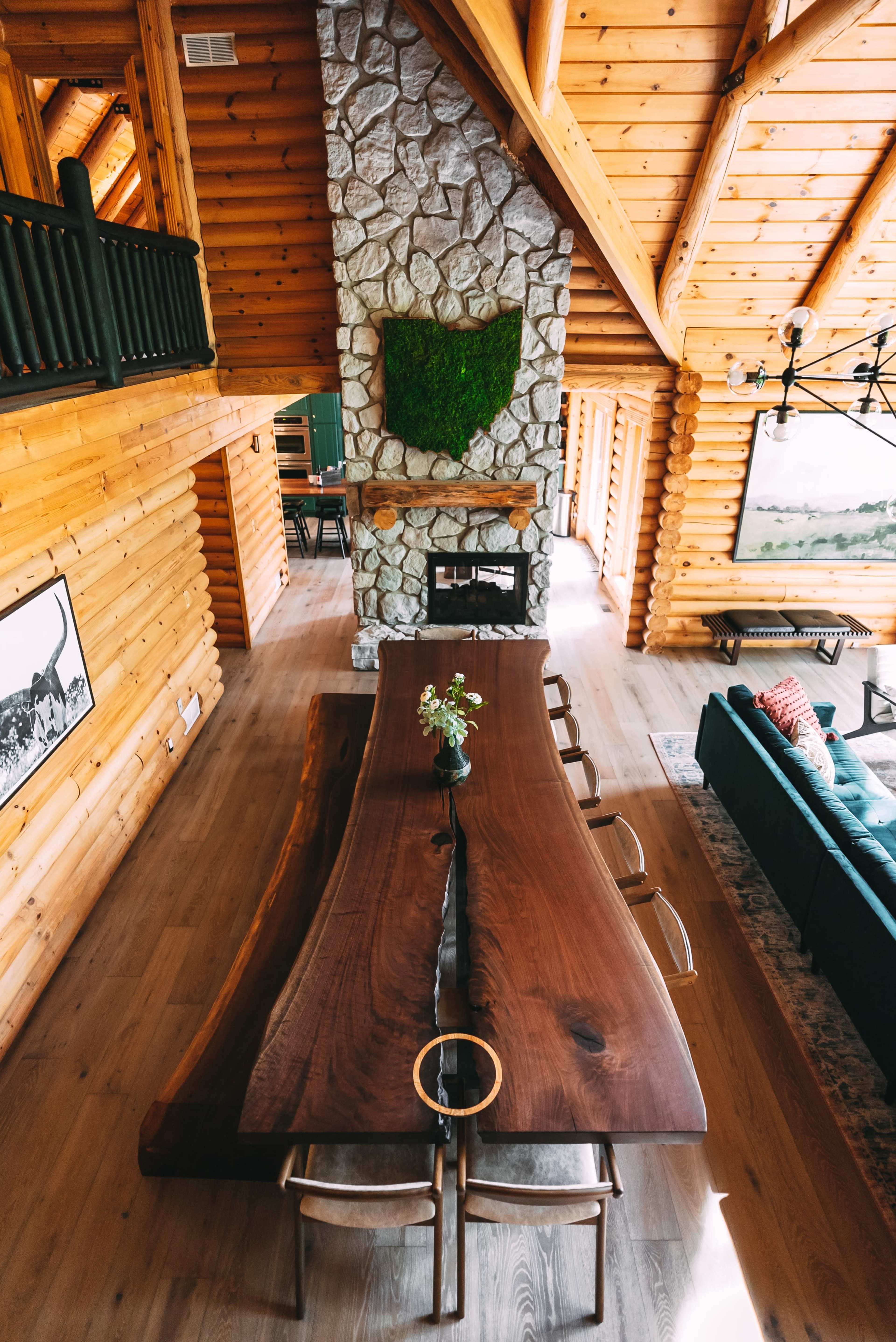 The image shows a spacious wooden cabin interior featuring a long rustic table, a stone fireplace, and a vaulted ceiling.