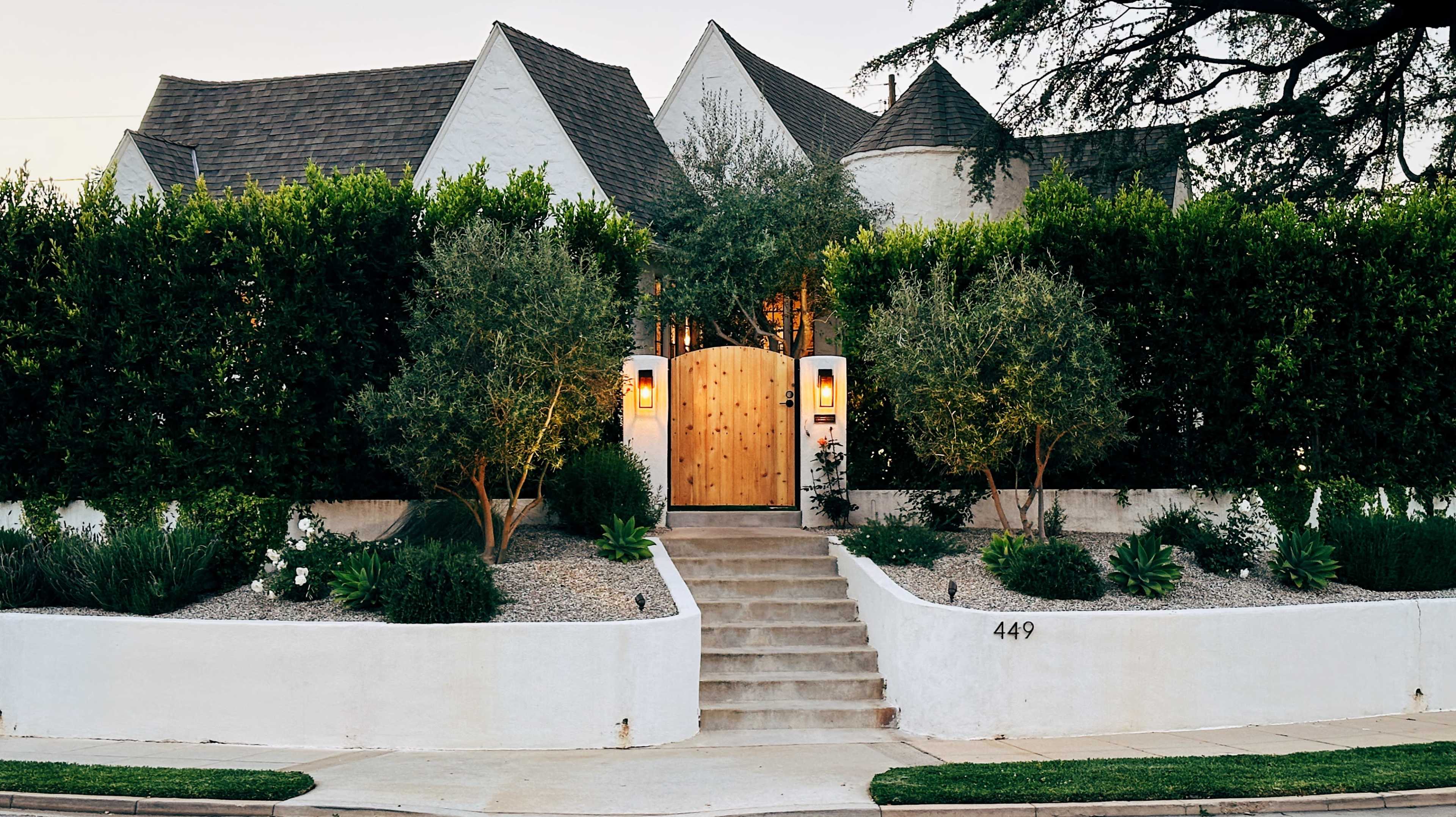 A modern house with a wooden gate surrounded by a landscaped garden and a stone pathway is set against a backdrop of tall hedges.