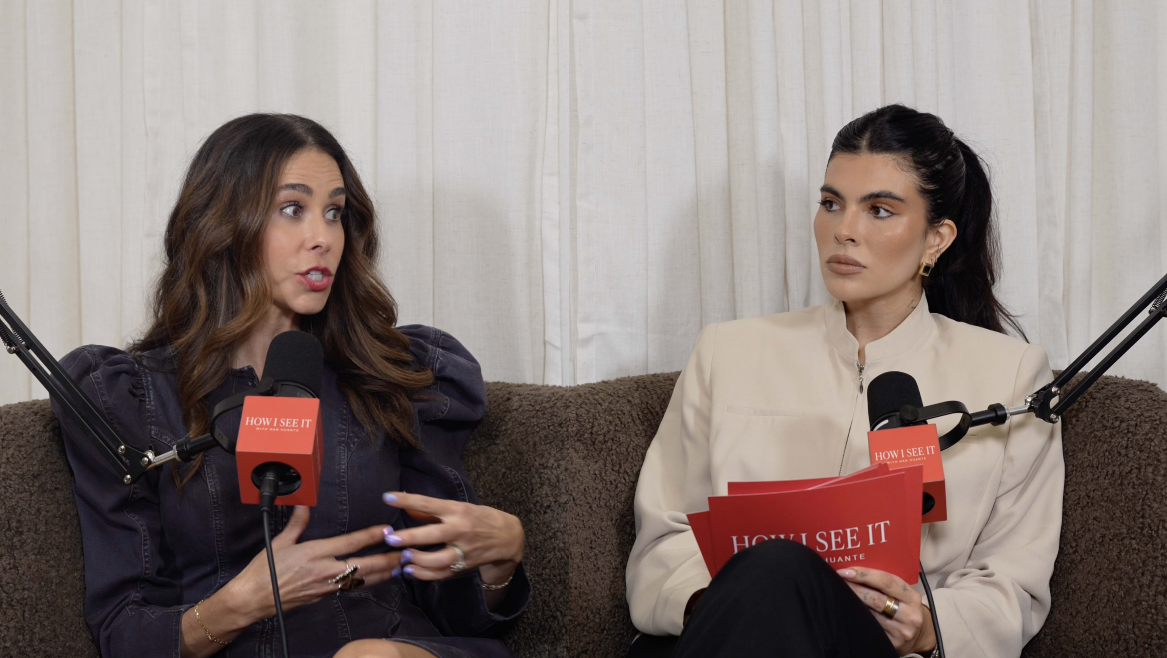 Two women are seated on a couch, discussing while holding red signs with the text "HOW I SEE IT" and microphones positioned in front of them.