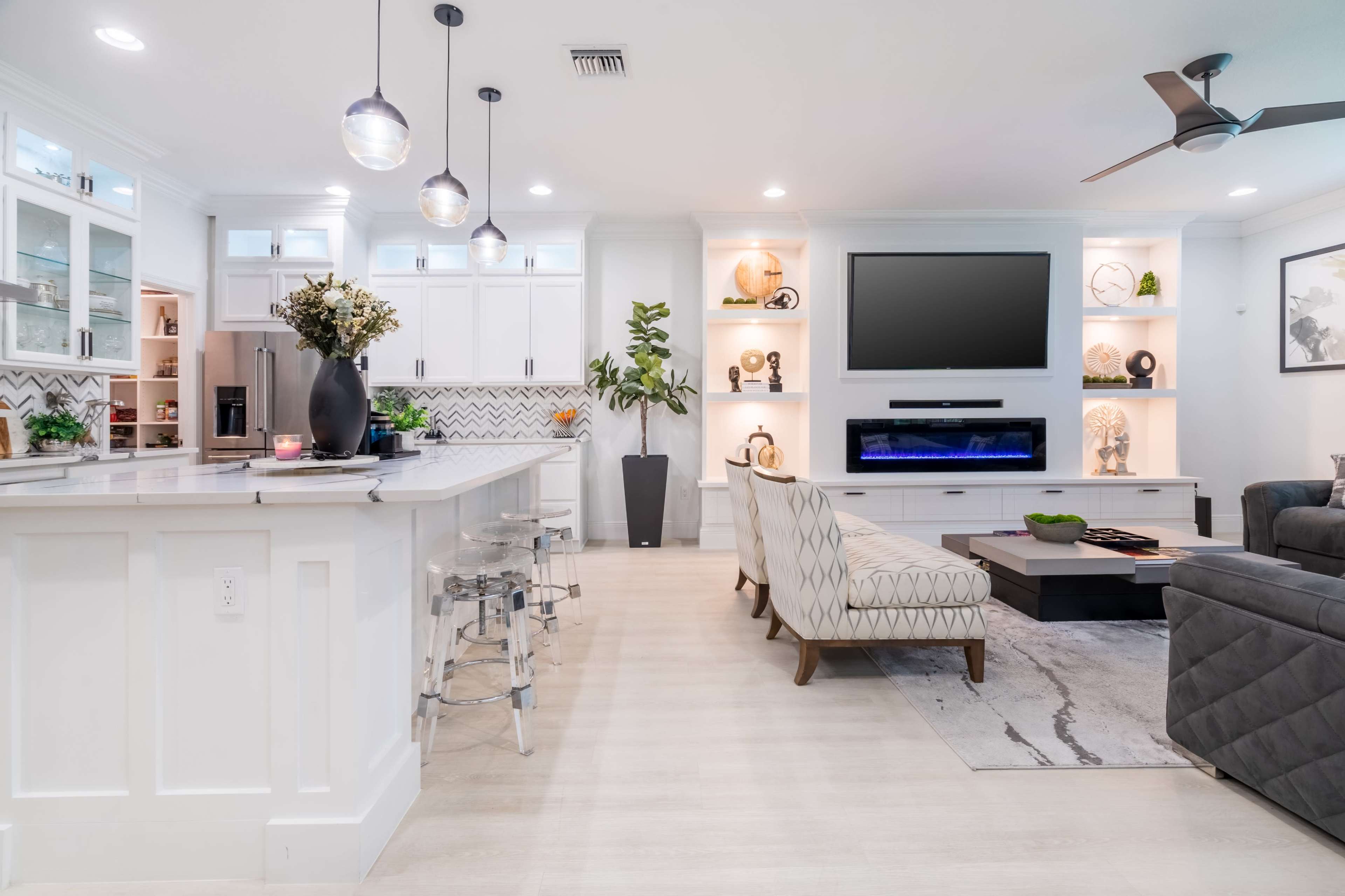 A modern living room with a light-colored floor, a flat-screen TV mounted on the wall, and a kitchen area featuring white cabinetry and bar seating.
